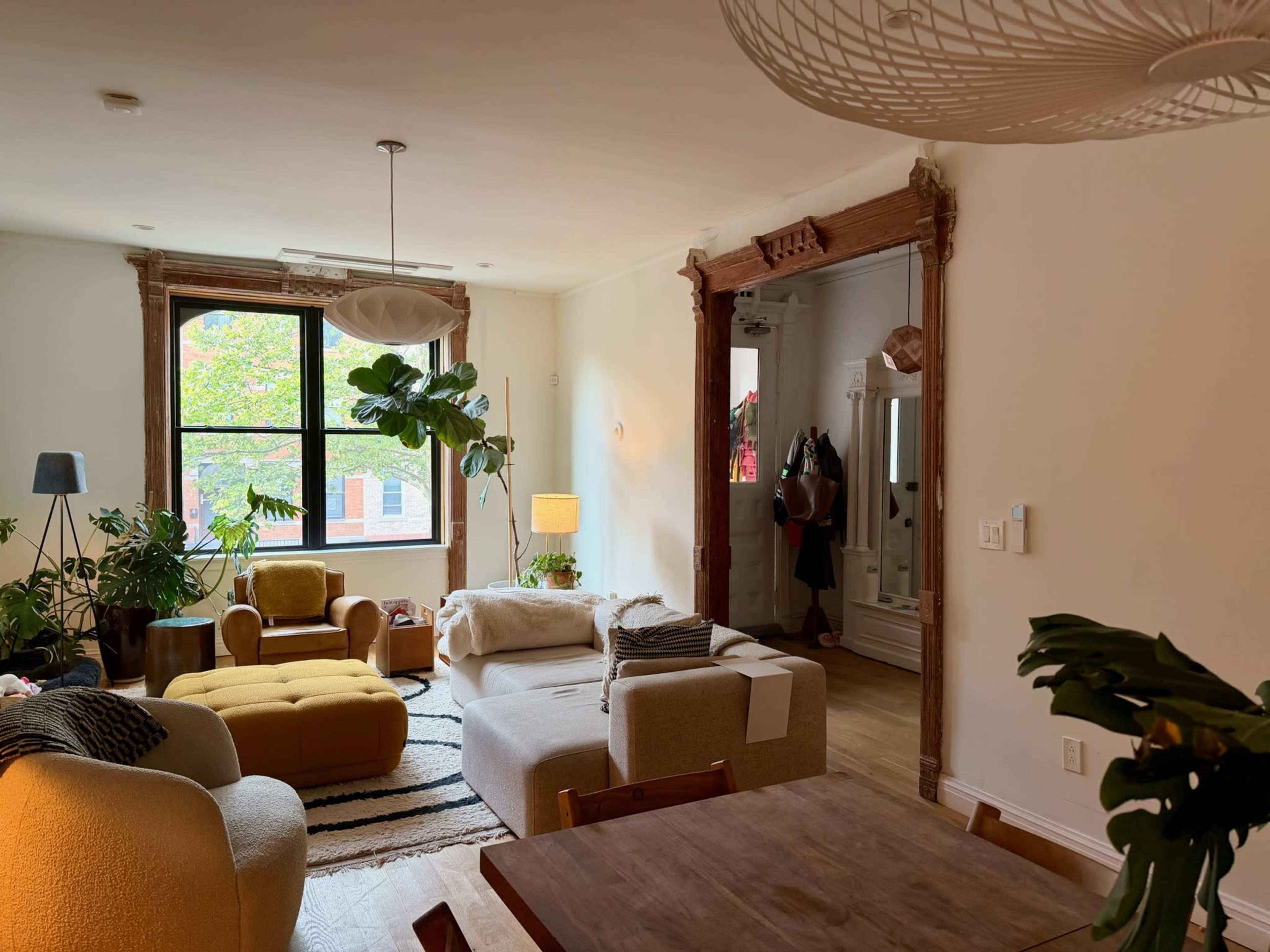 The image shows a modern living room with plants, comfortable seating, and a dining area, featuring natural light from large windows.