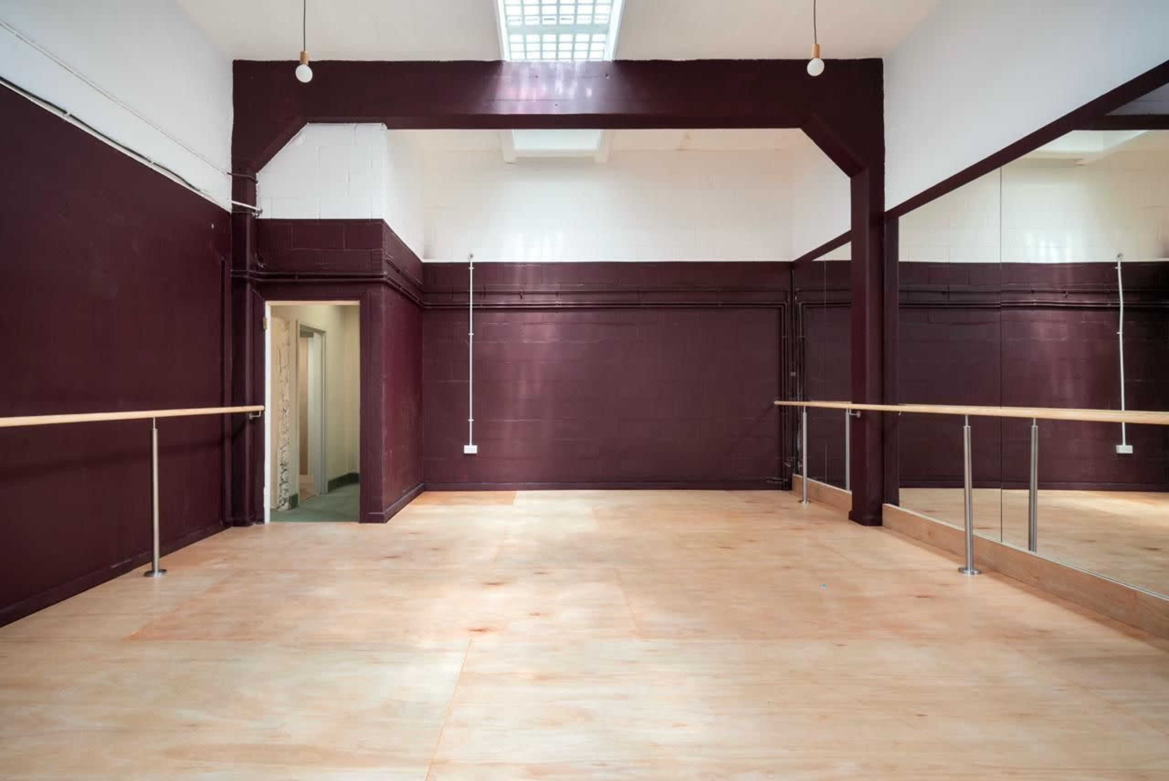 The image shows an empty dance studio with a wooden floor, mirrored wall, and a large overhead skylight.