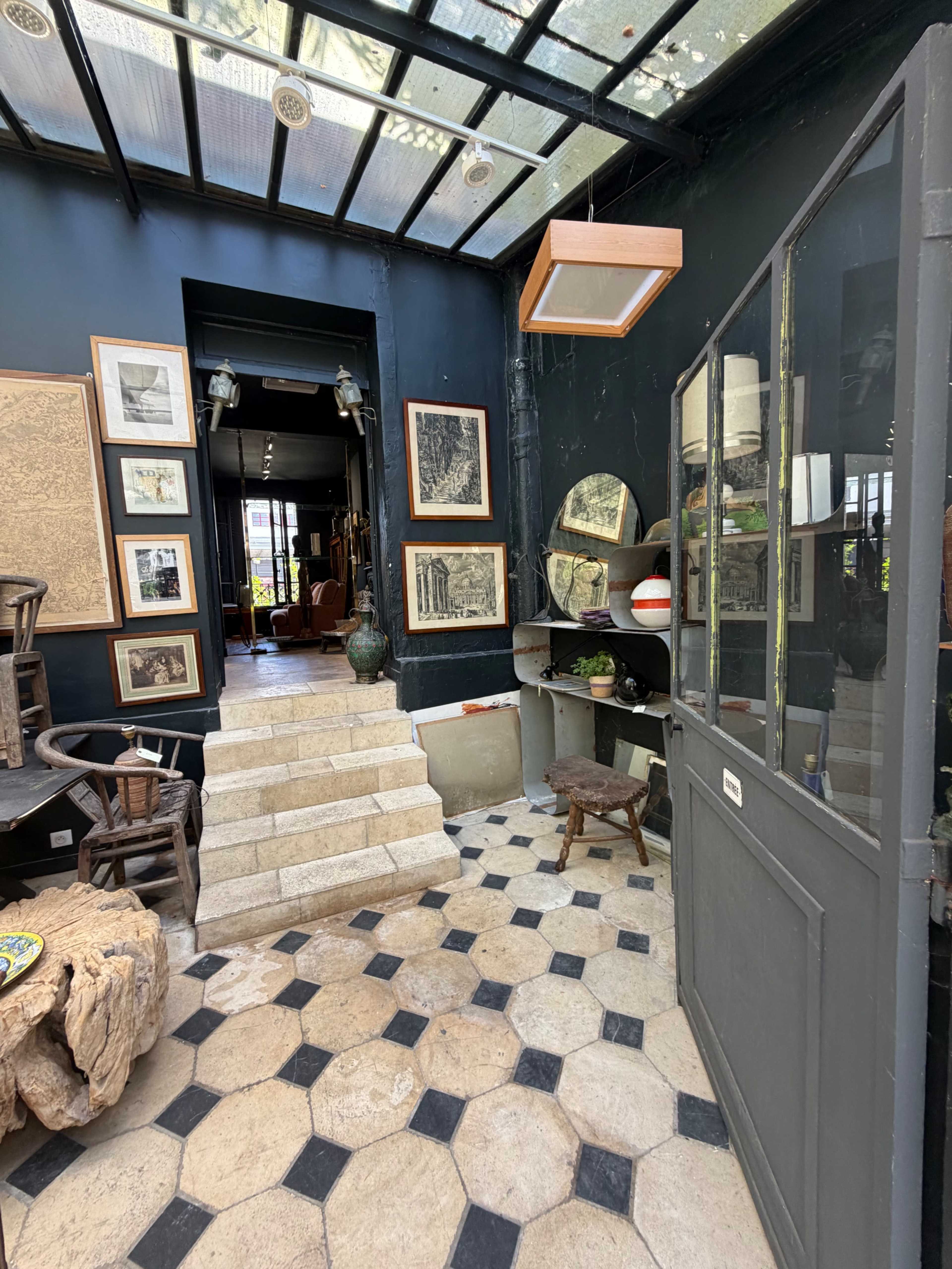 The image shows a stylish entrance area with a glass ceiling, dark walls adorned with framed artwork, and a staircase leading to another room.