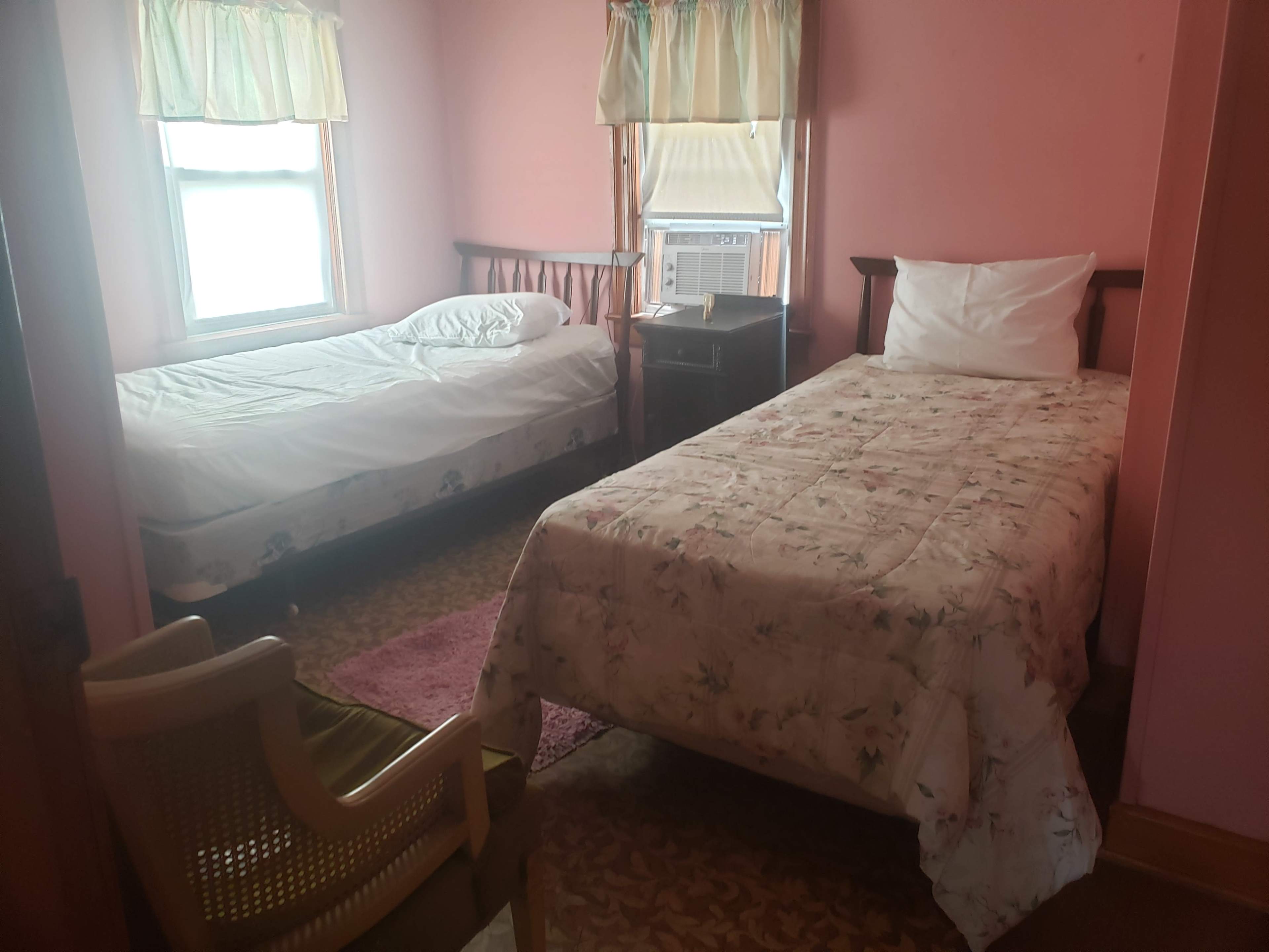 A small, pink-painted bedroom with two beds, an air conditioning unit, and window with light curtains.