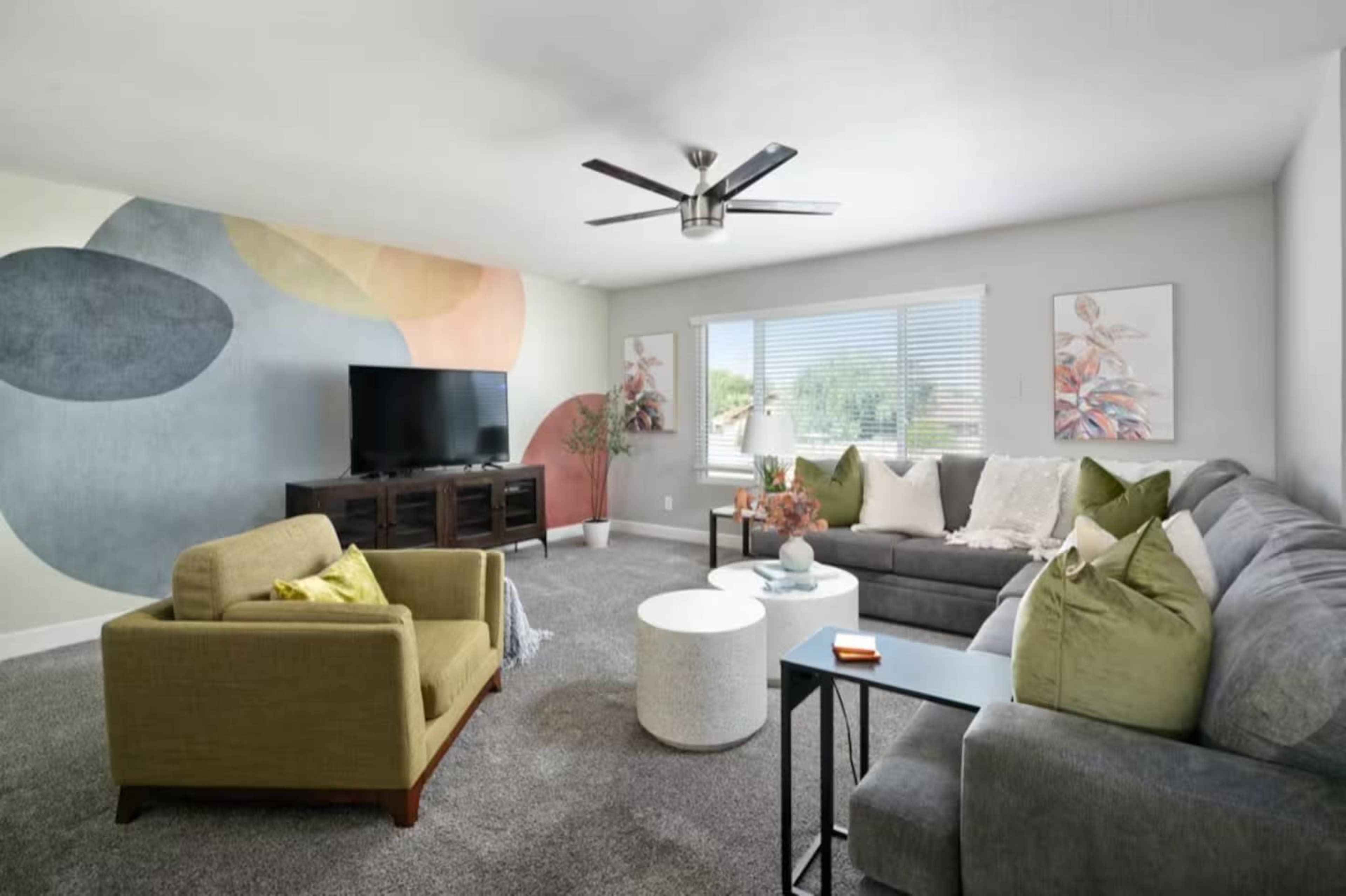 The living room features a gray sofa, a green armchair, a white circular coffee table, a television on a wooden stand, and a colorful abstract mural on one wall.