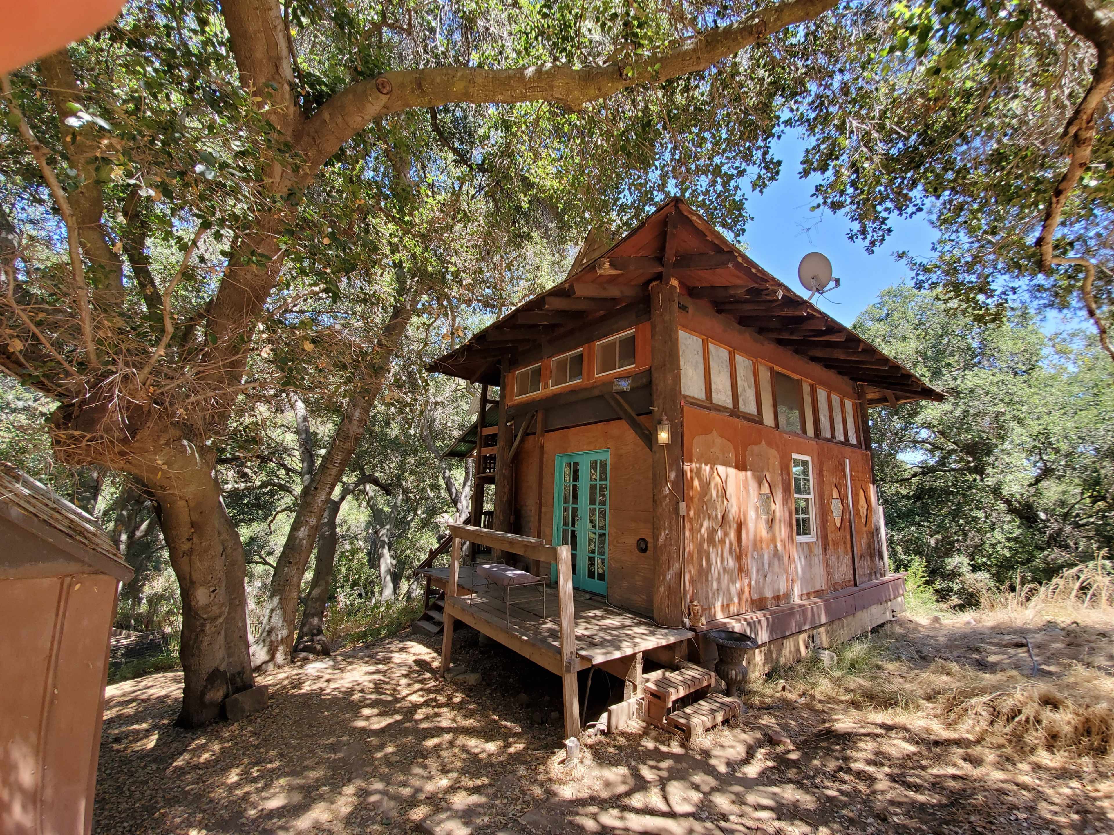 A small wooden house with a porch, situated among large trees in a sunny clearing.