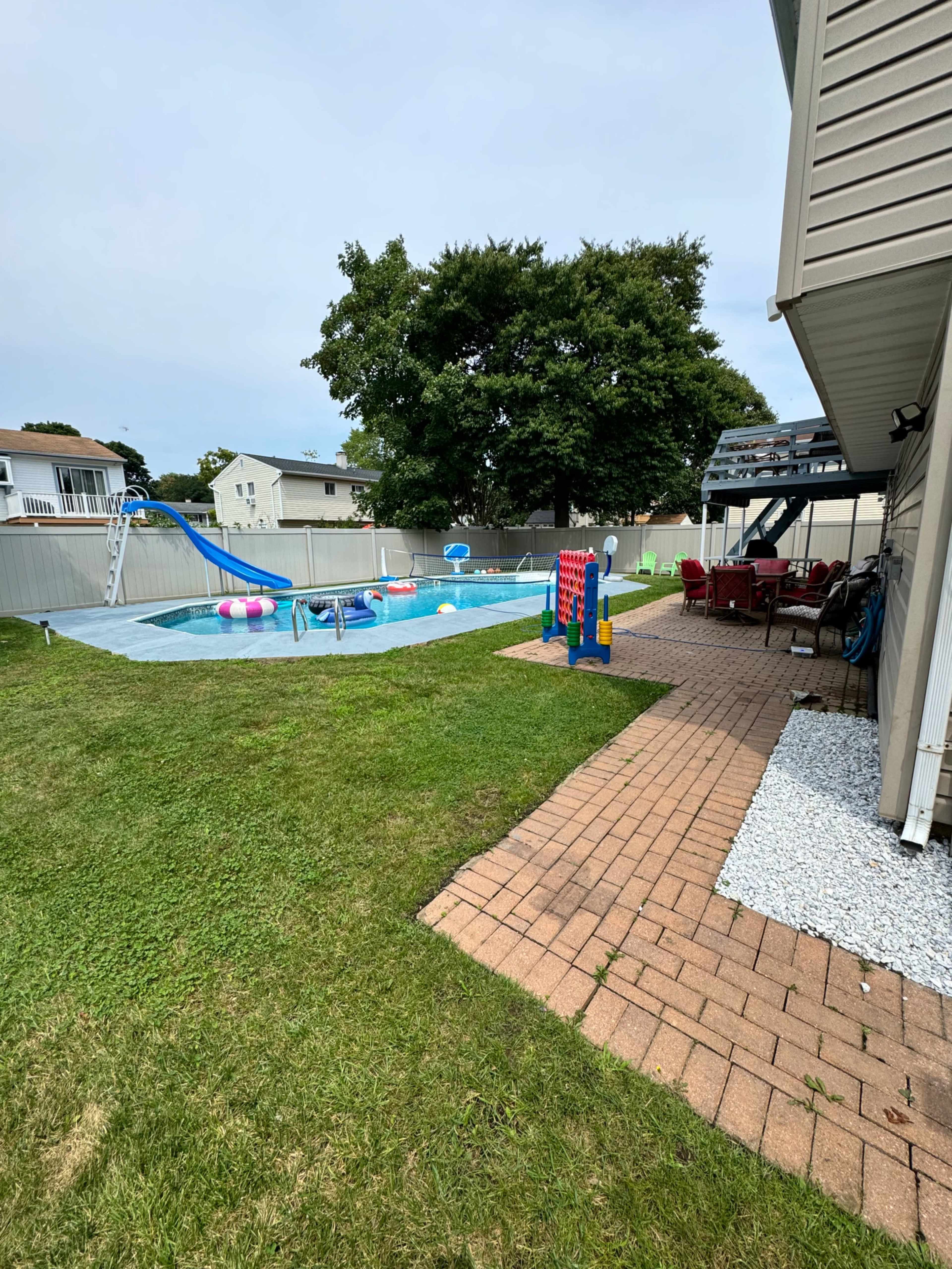 A residential backyard features a swimming pool with a slide, surrounded by a patio area and green lawn.