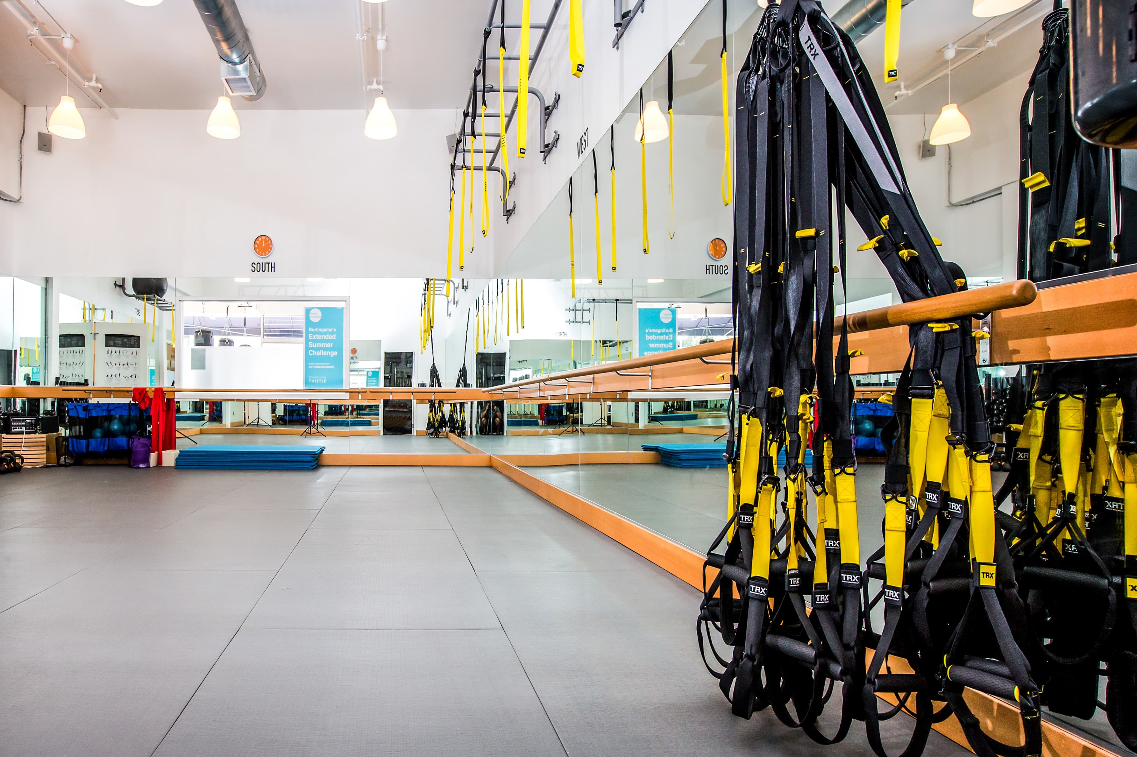 A gym interior equipped with suspension training straps hanging from a mirrored wall, with workout mats and exercise equipment in the background.