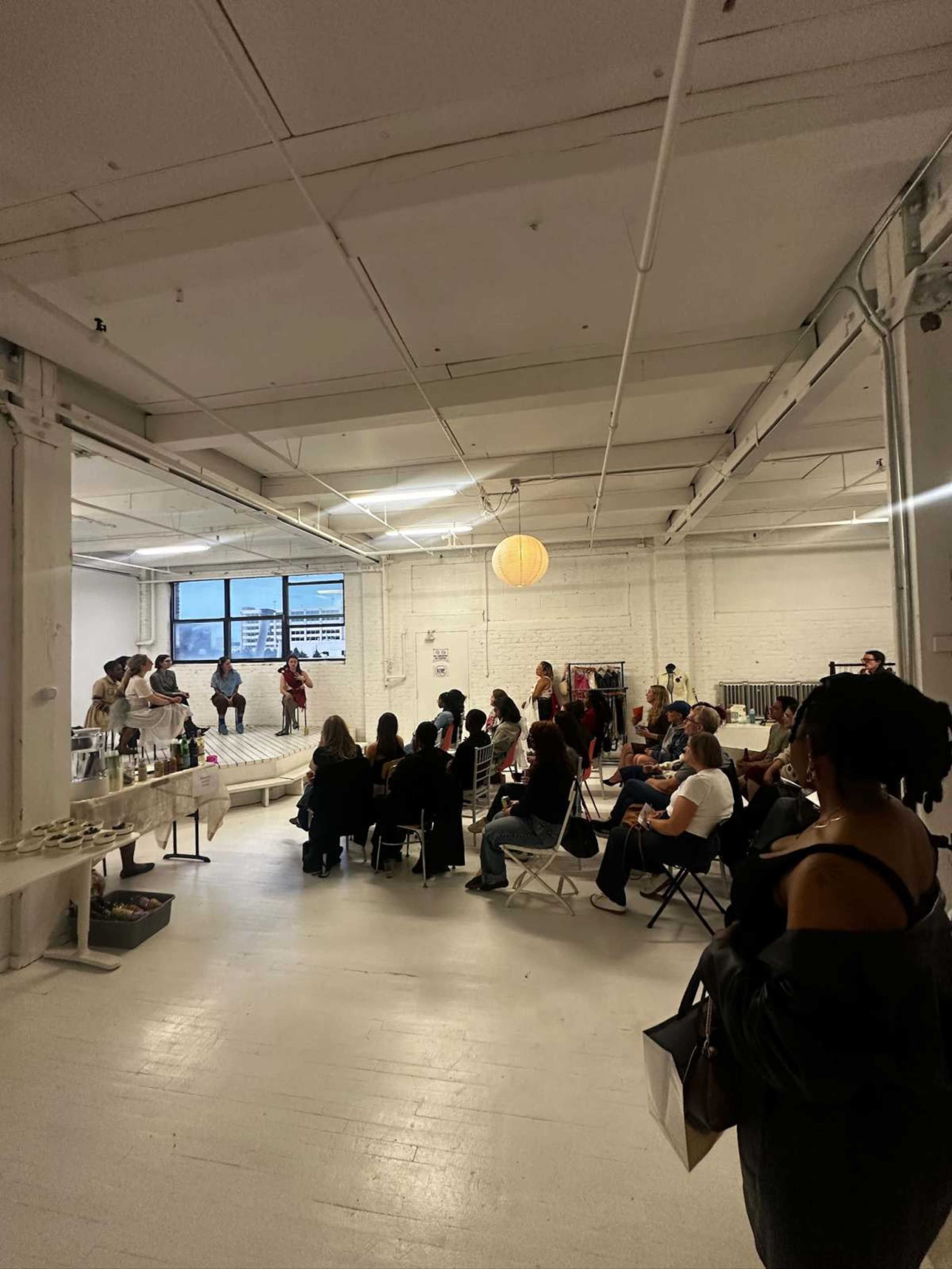 A gathering is taking place in a spacious, well-lit room with seated attendees facing a small stage or presentation area.