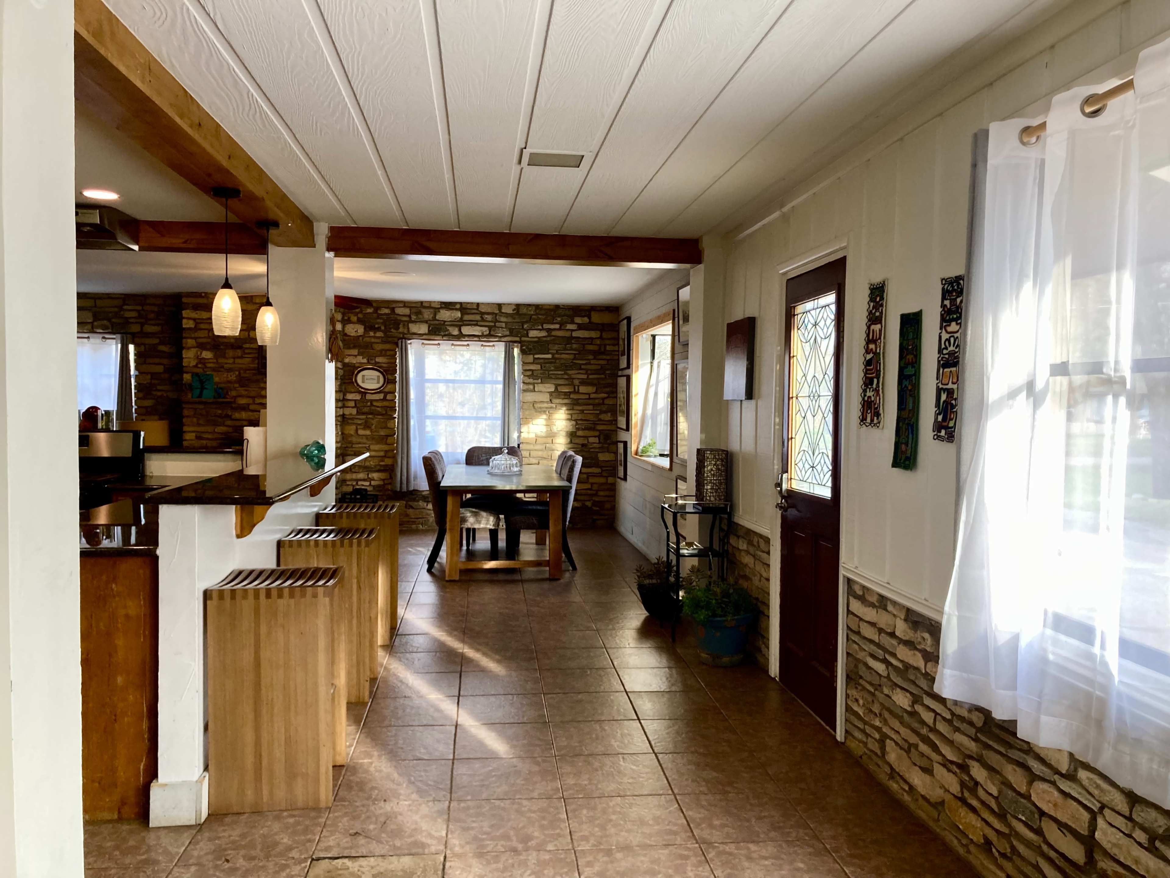 A spacious interior with a dining area featuring a round table and chairs, and a kitchen bar with wooden stools, all set against a backdrop of stone walls and natural light from windows.