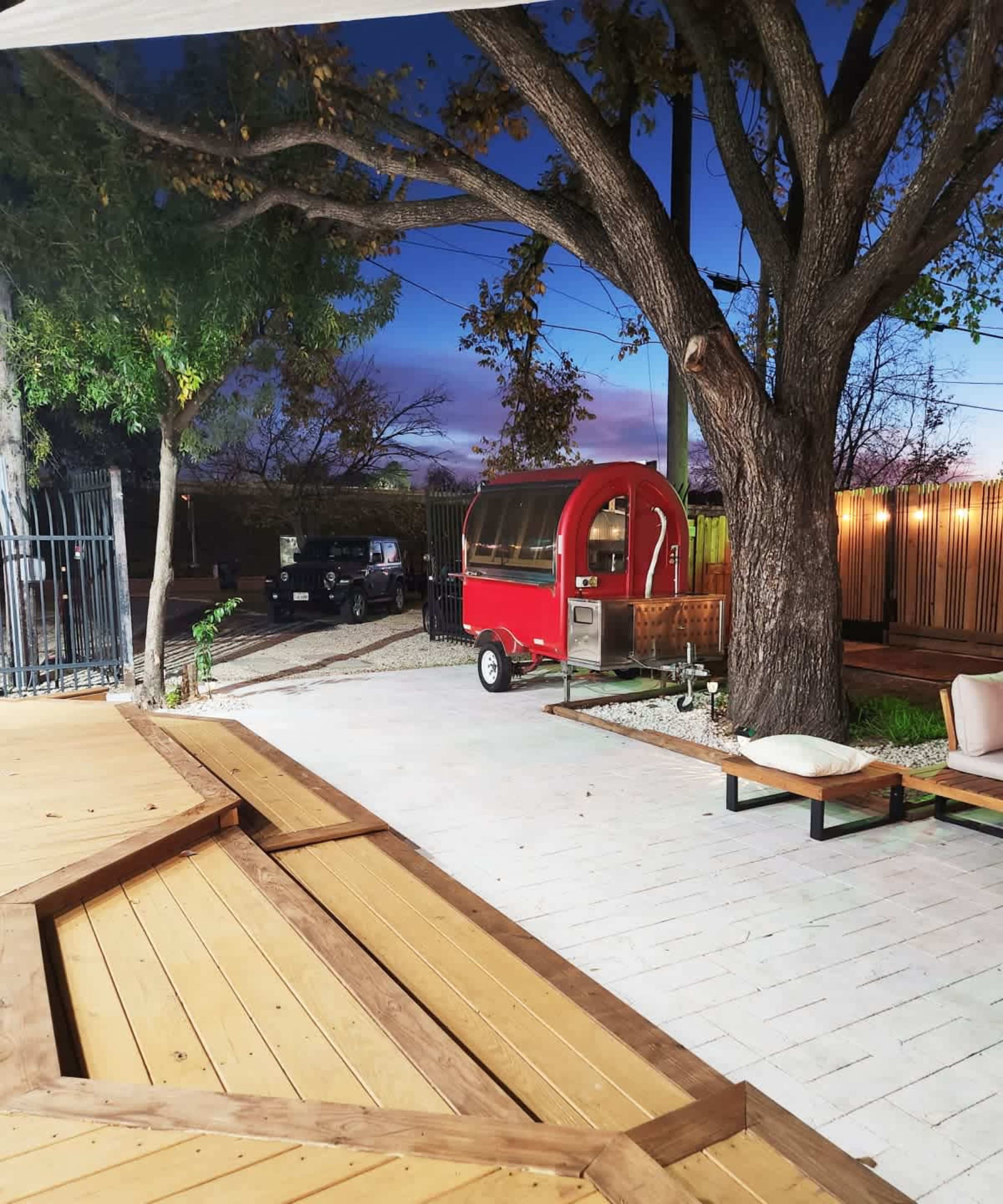The image shows a red food truck parked on a gravel path beside a wooden deck and tree, with a dusk sky in the background.