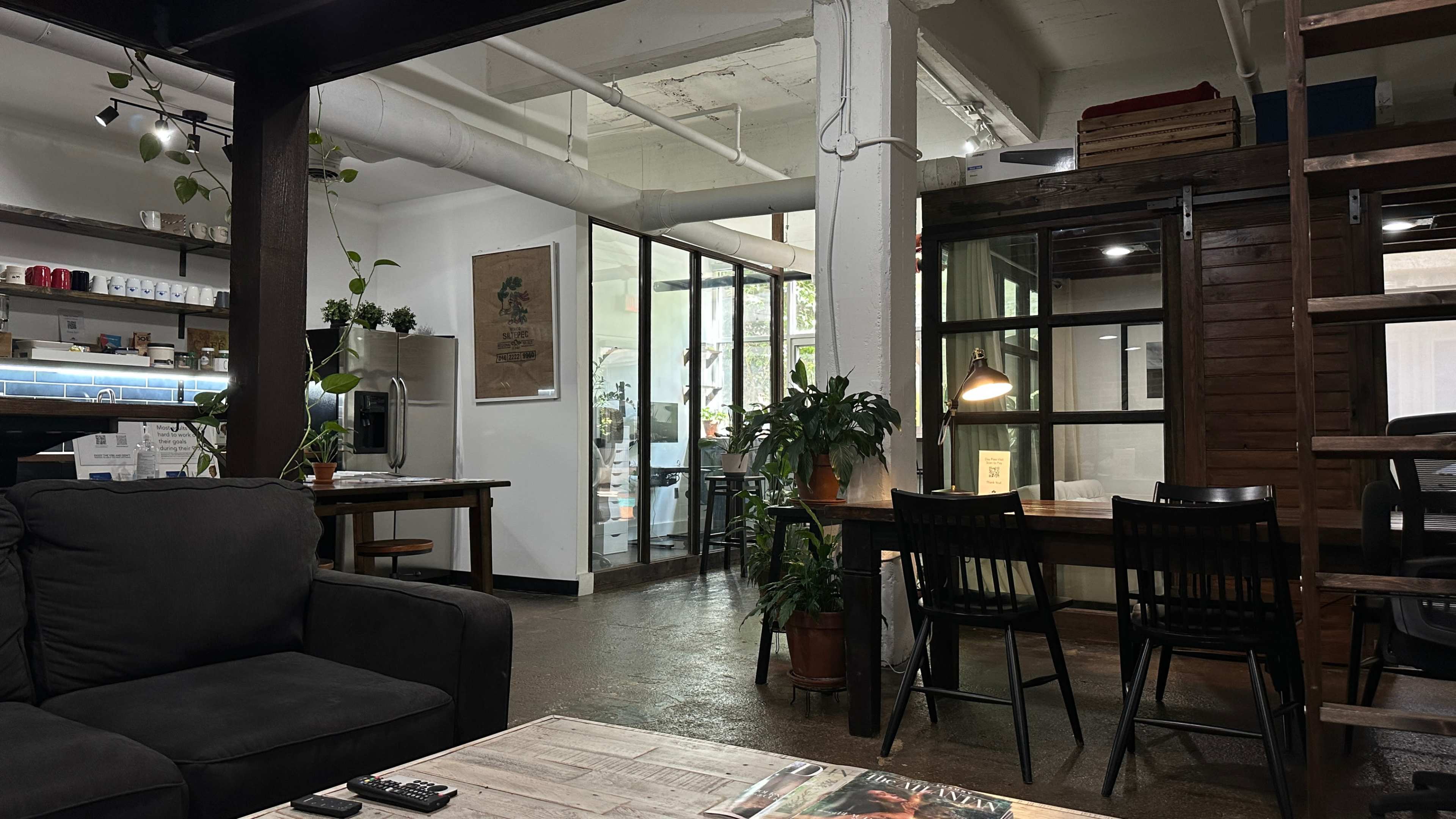 The image shows a modern, open workspace featuring a seating area with a couch, plants, and a glass-walled section that leads to a kitchen or a communal area.