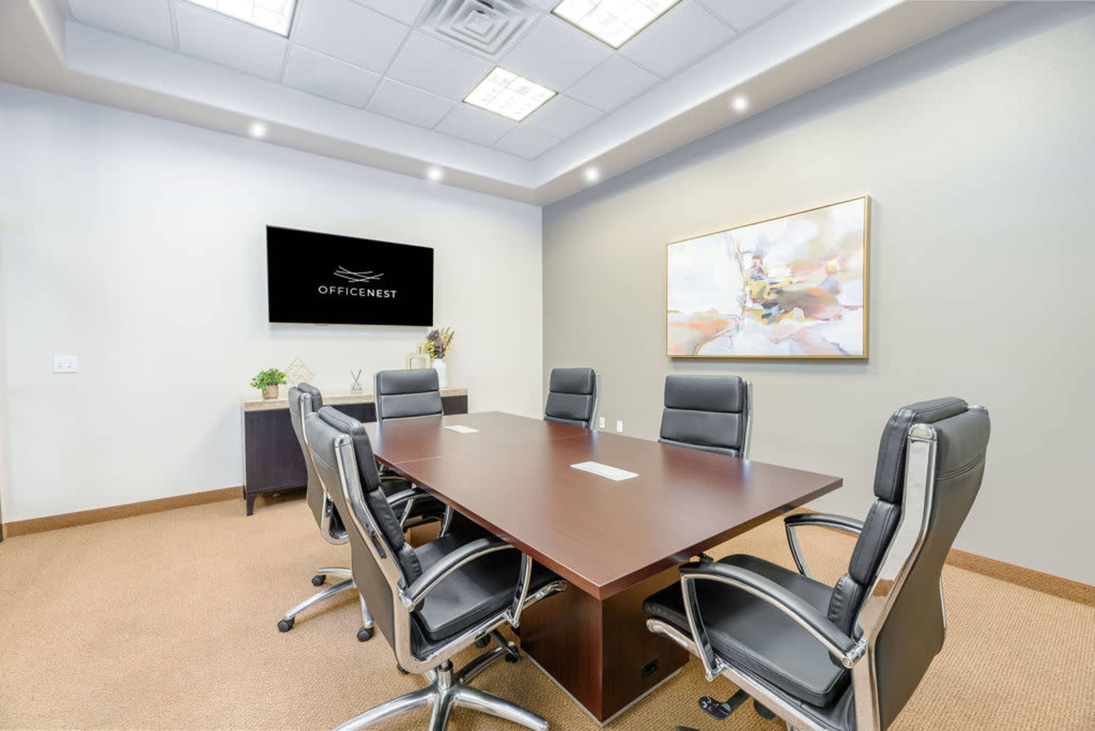 A modern conference room features a large wooden table surrounded by six black leather chairs, with a wall-mounted screen and a decorative artwork.