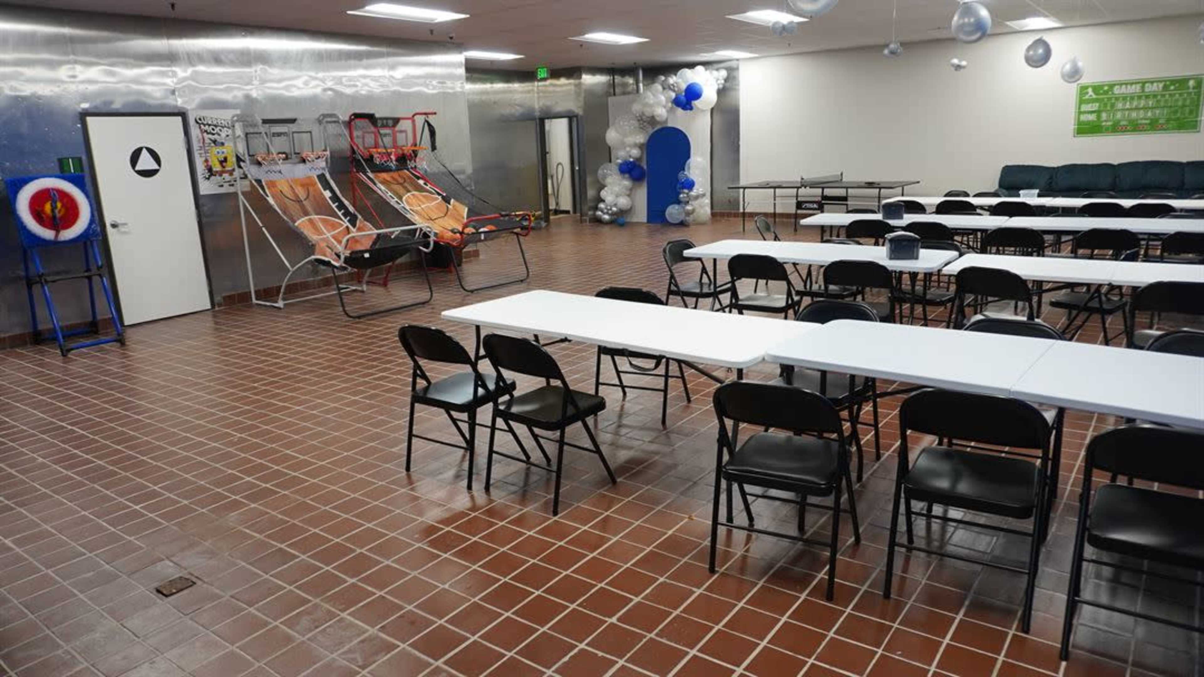 The image shows a large room with two rows of folding tables set up for an event, surrounded by chairs, and featuring a silver wall with decorative elements including a blue arch and several outdoor lounge chairs.