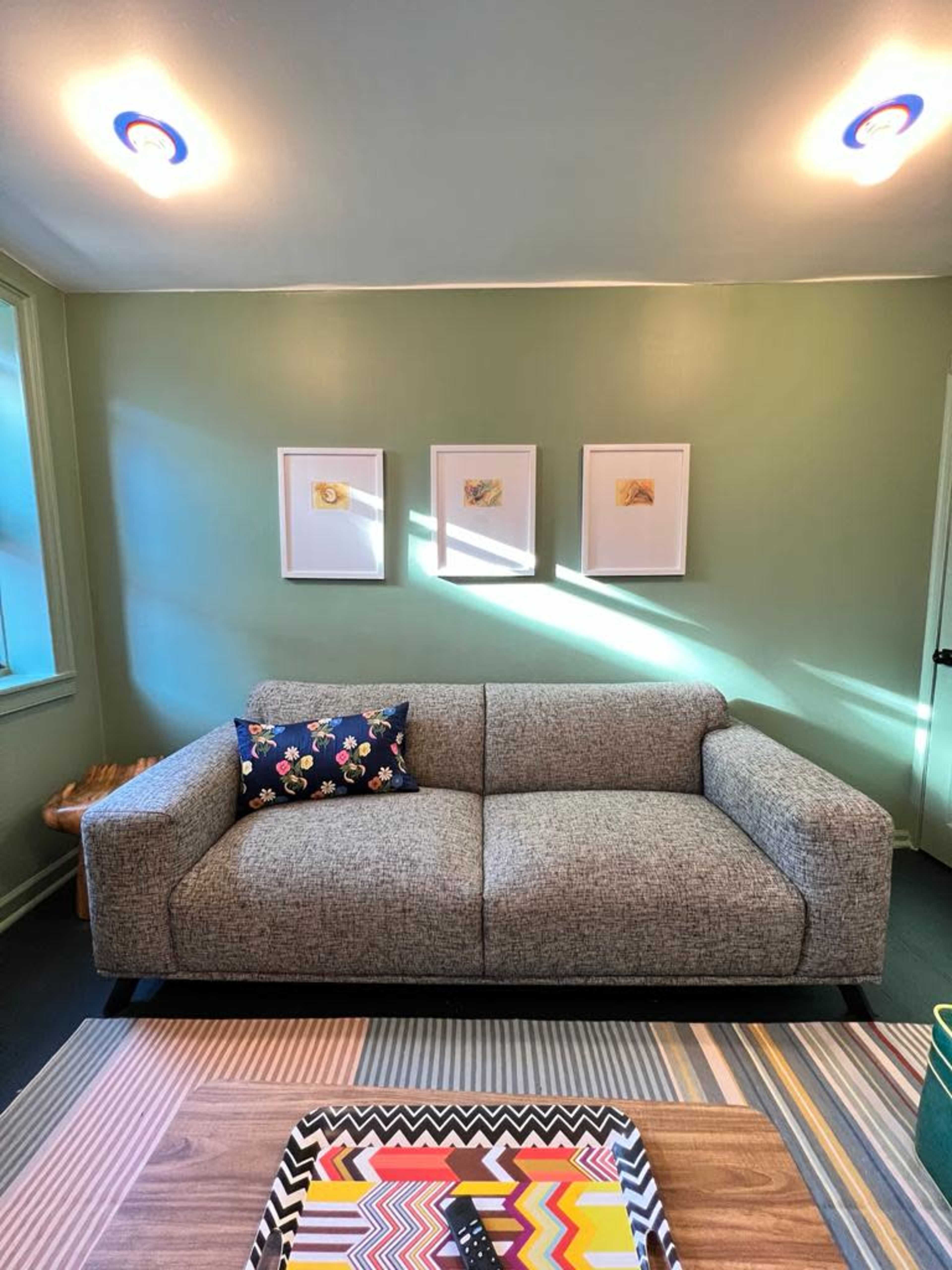 A gray couch with a patterned pillow is positioned against a green wall, flanked by three framed artworks, with light streaming in from the windows.