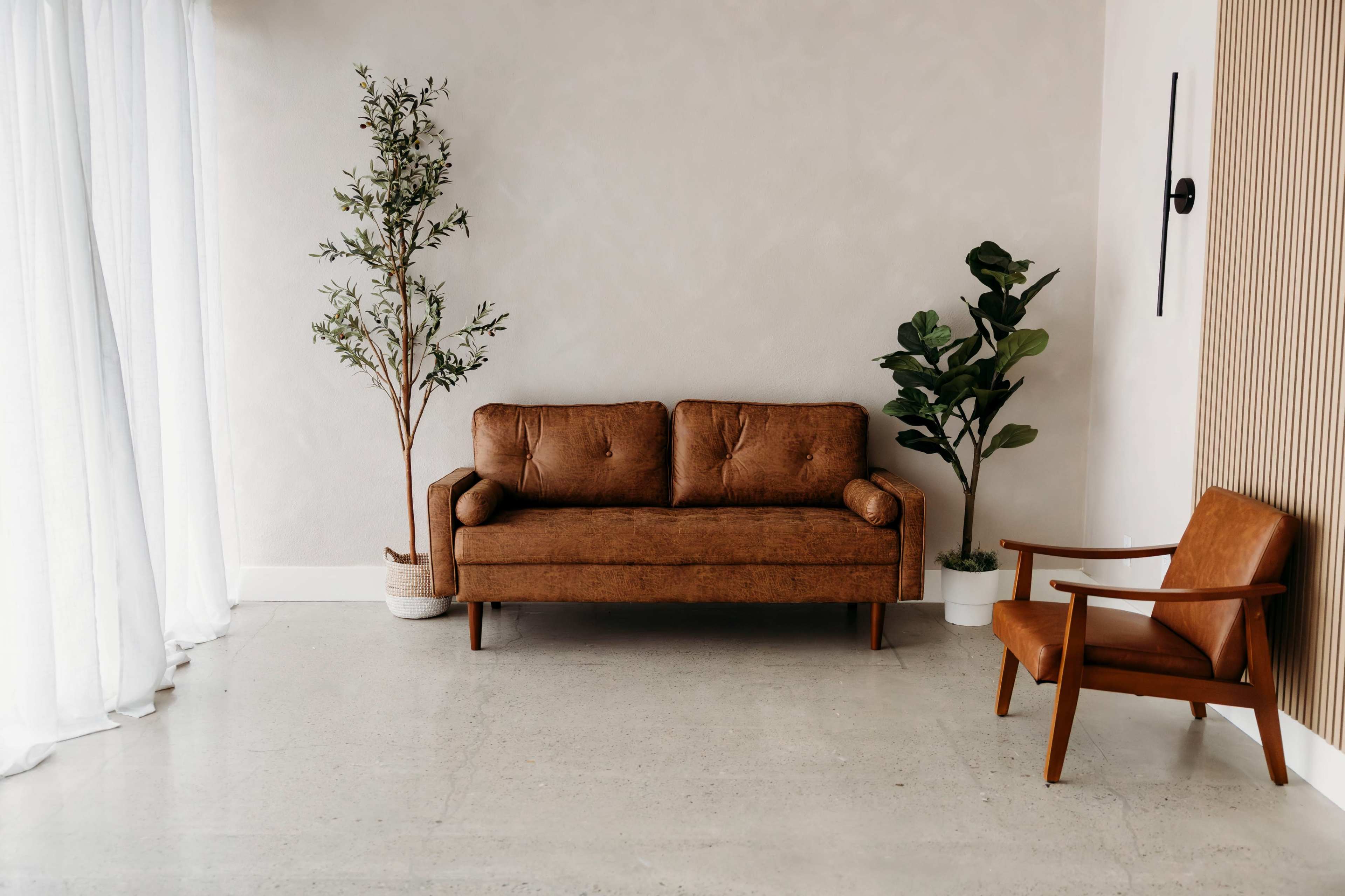 The image shows a minimalist interior featuring a brown sofa, a brown armchair, and two potted plants against a light-colored wall.