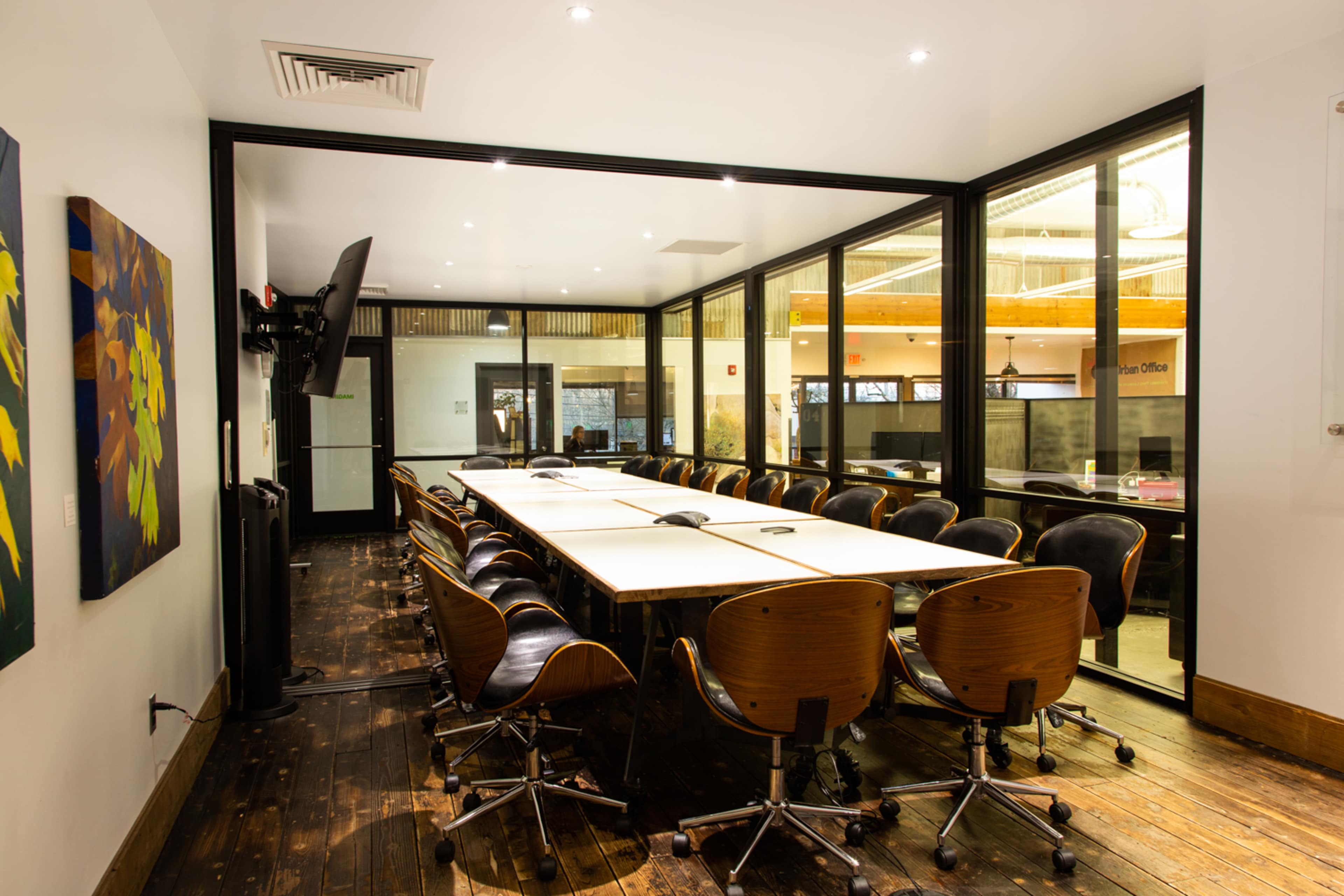 A modern conference room features a long wooden table surrounded by black chairs, with large glass walls and a television mounted on one side.