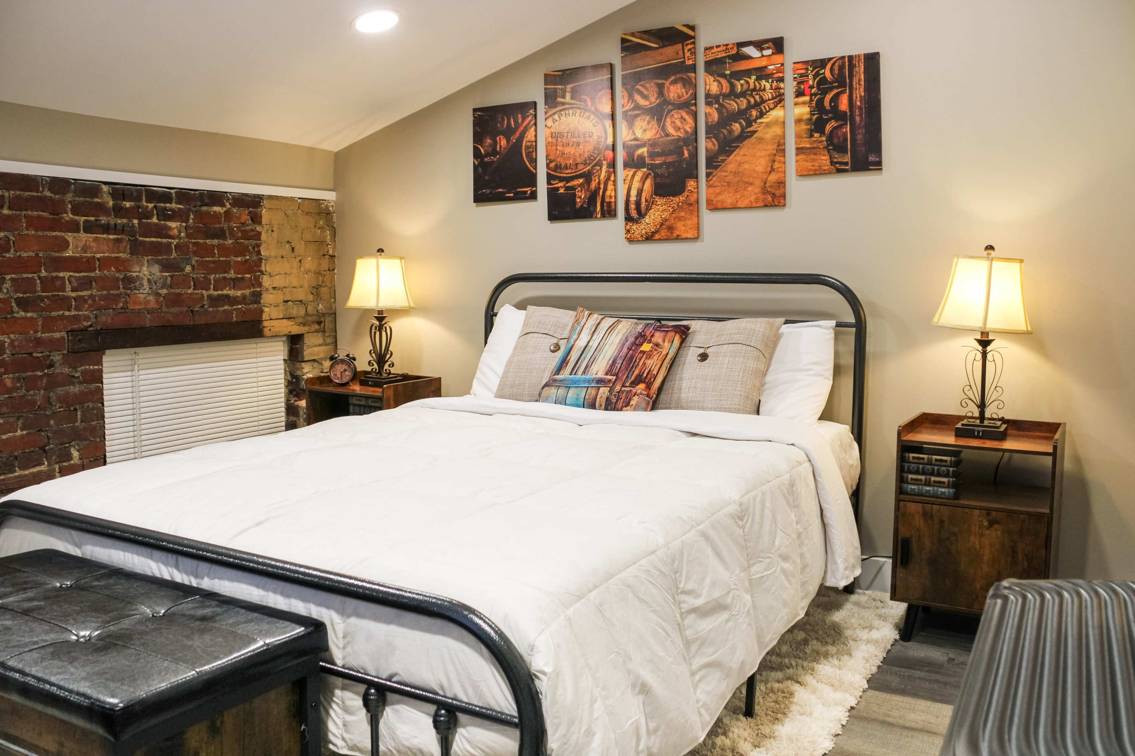 A cozy bedroom with a metal bed frame, a white comforter, and two bedside tables with lamps, complemented by exposed brick walls and artwork above the bed.