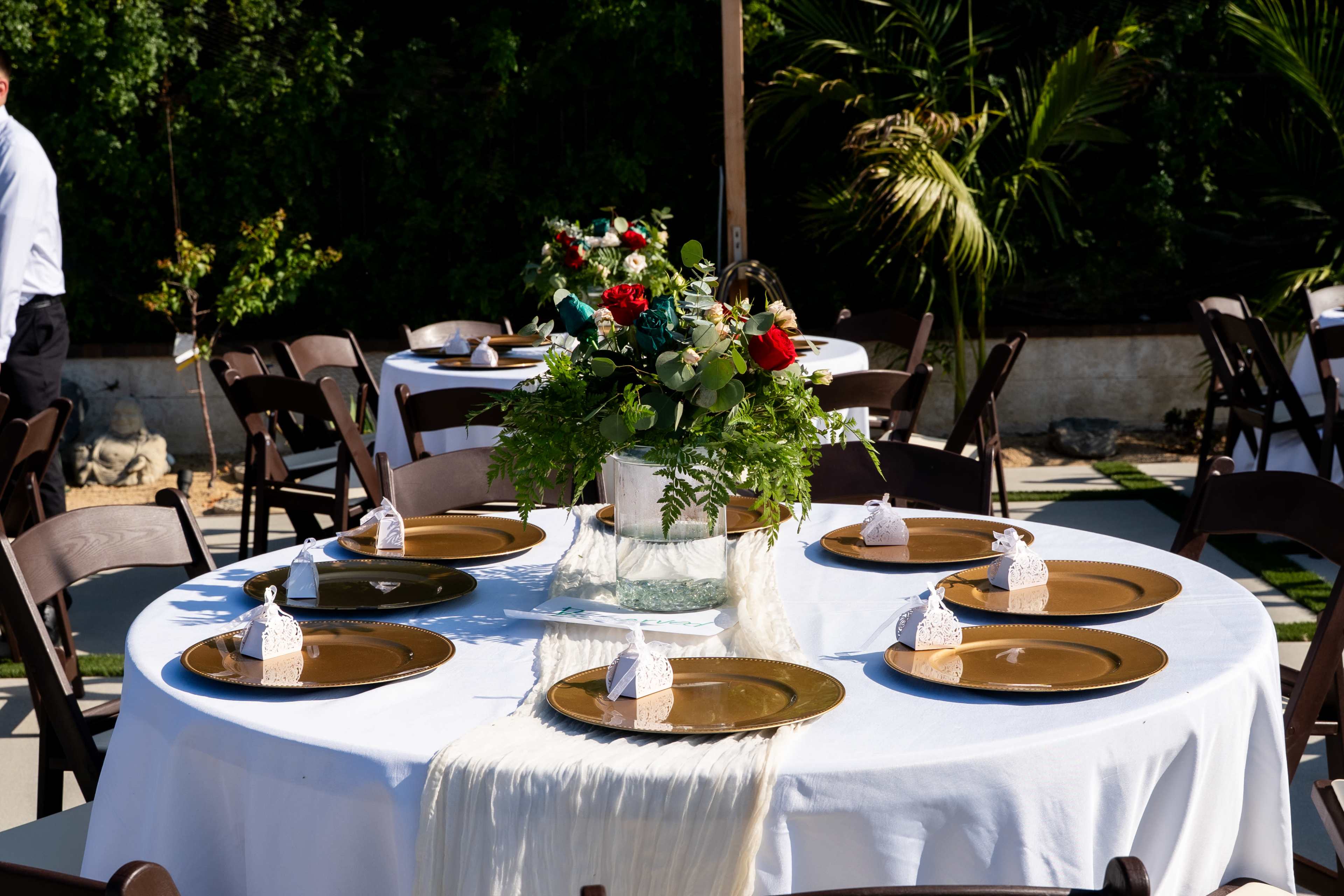 A round table is set for a meal with gold plates, white napkins shaped like little hats, and a floral centerpiece surrounded by green foliage.