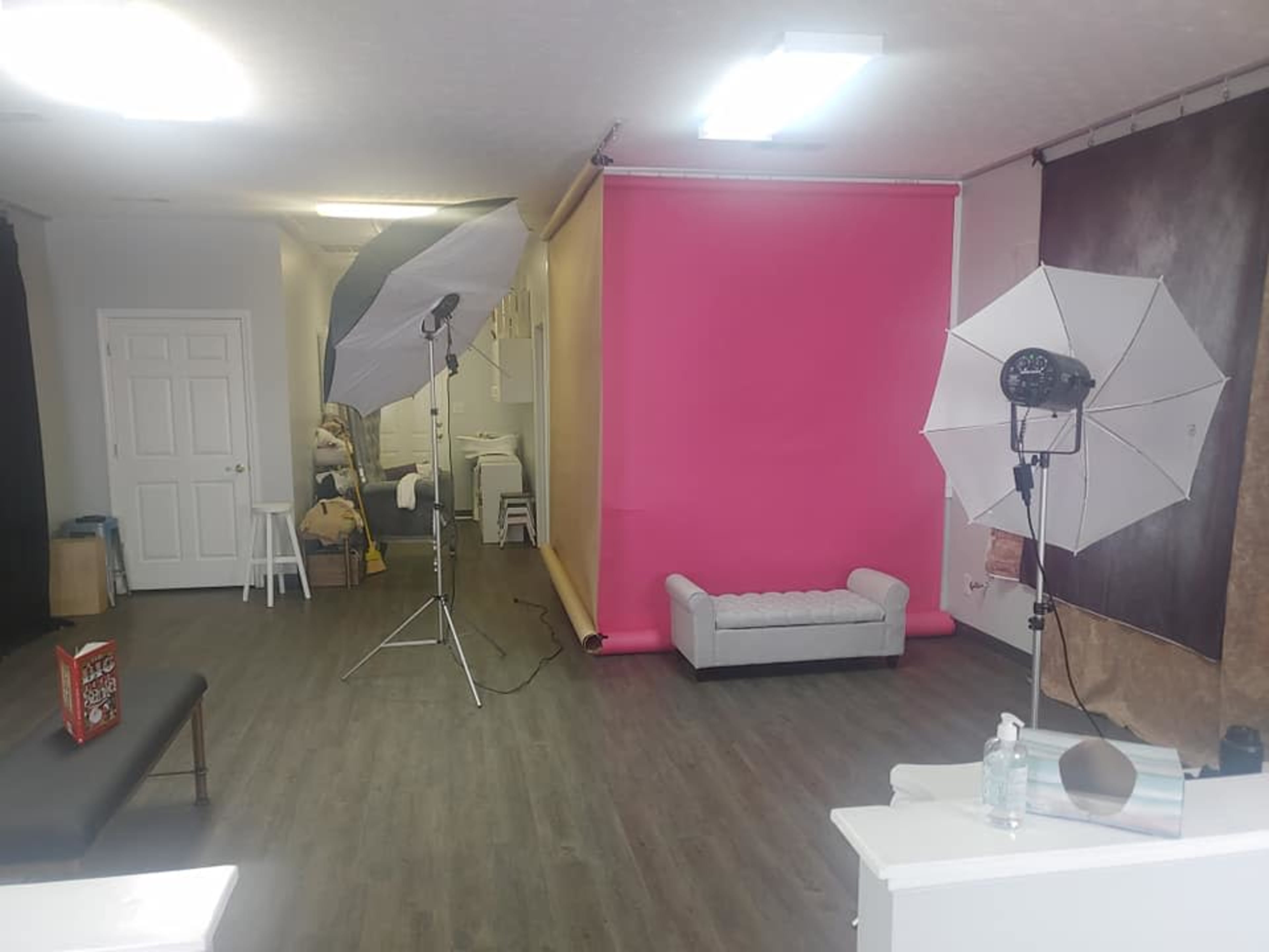 A photo studio with a pink backdrop, two softbox lights, a gray sofa, and various photography equipment arranged throughout the space.