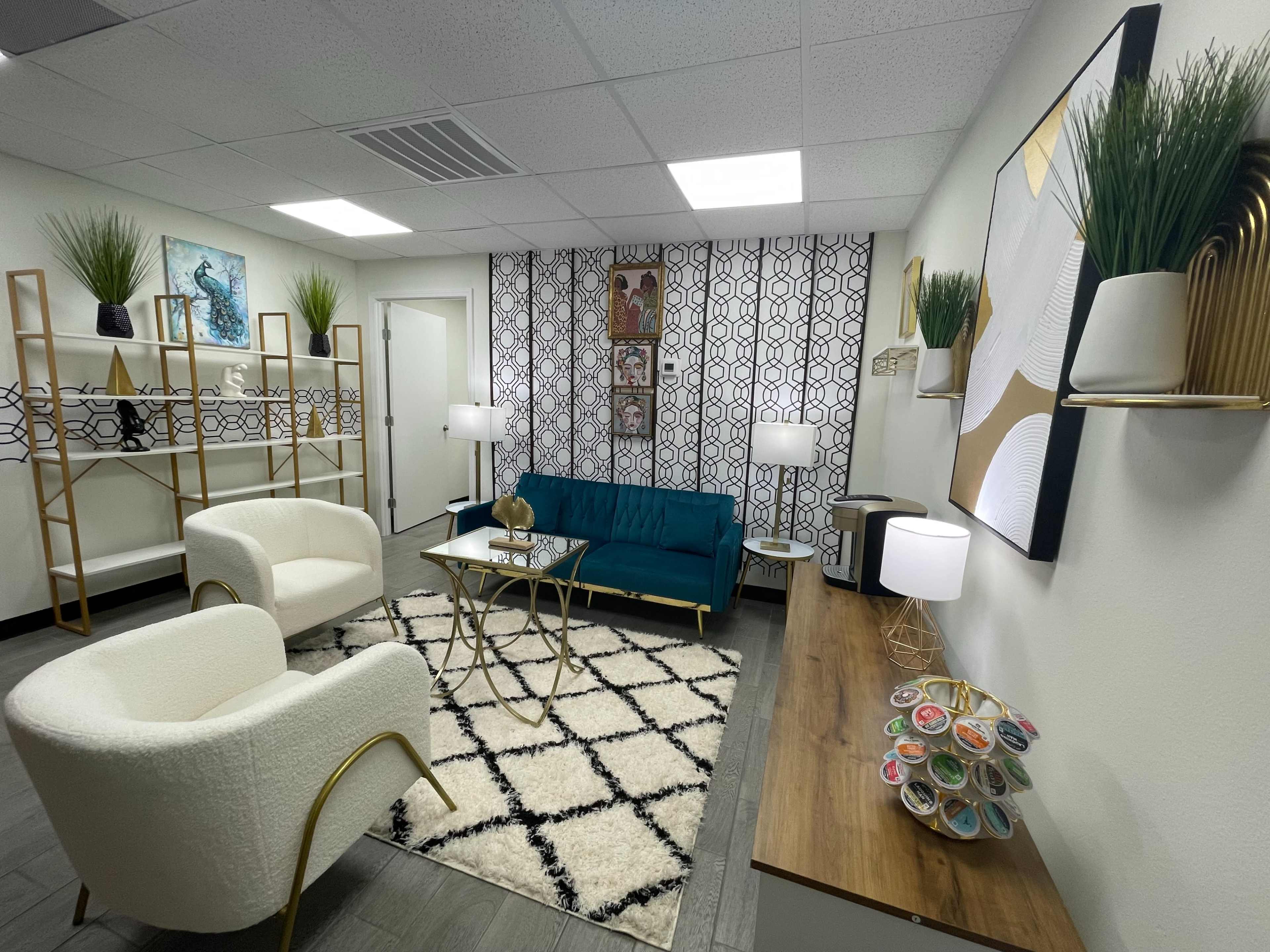 The image shows a modern lounge area with a teal sofa, two white armchairs, a coffee table, and decorative plants on shelves against a patterned wall.