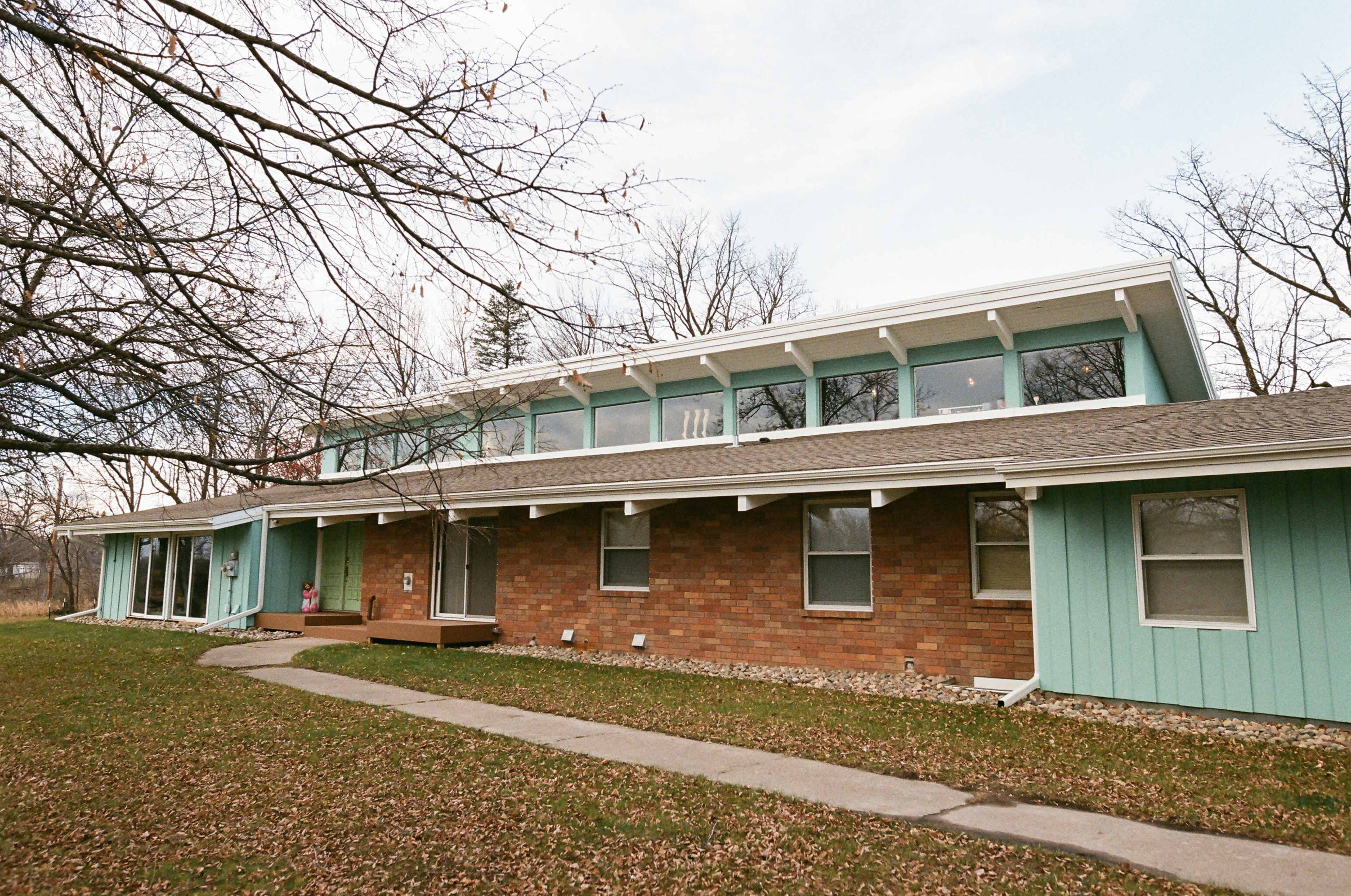 A large, two-story house with a combination of brick and turquoise siding sits on a grassy area surrounded by bare trees.