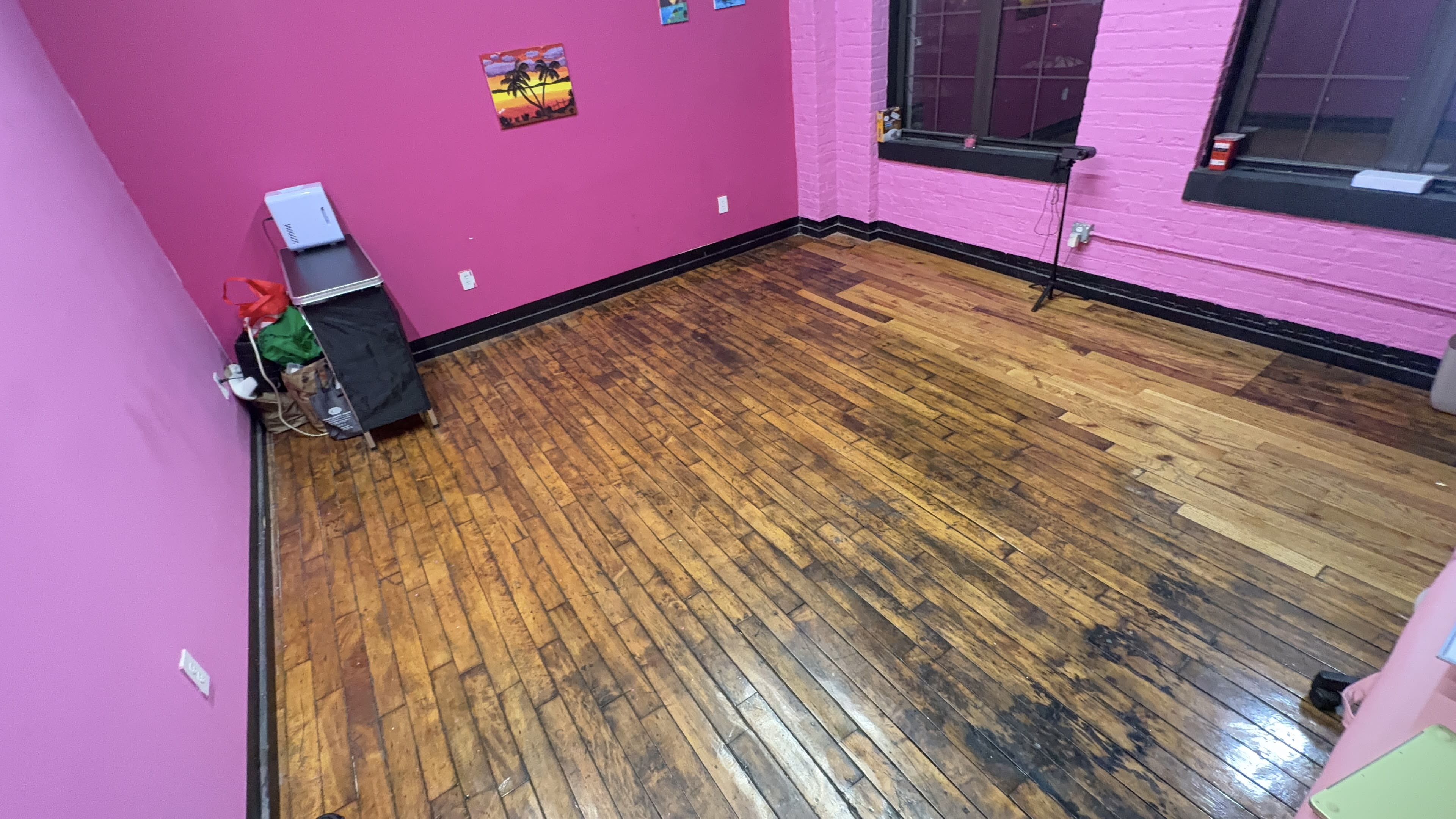 The image shows a room with pink walls, wooden flooring, and minimal furniture, including a metal cart and some artwork on the walls.