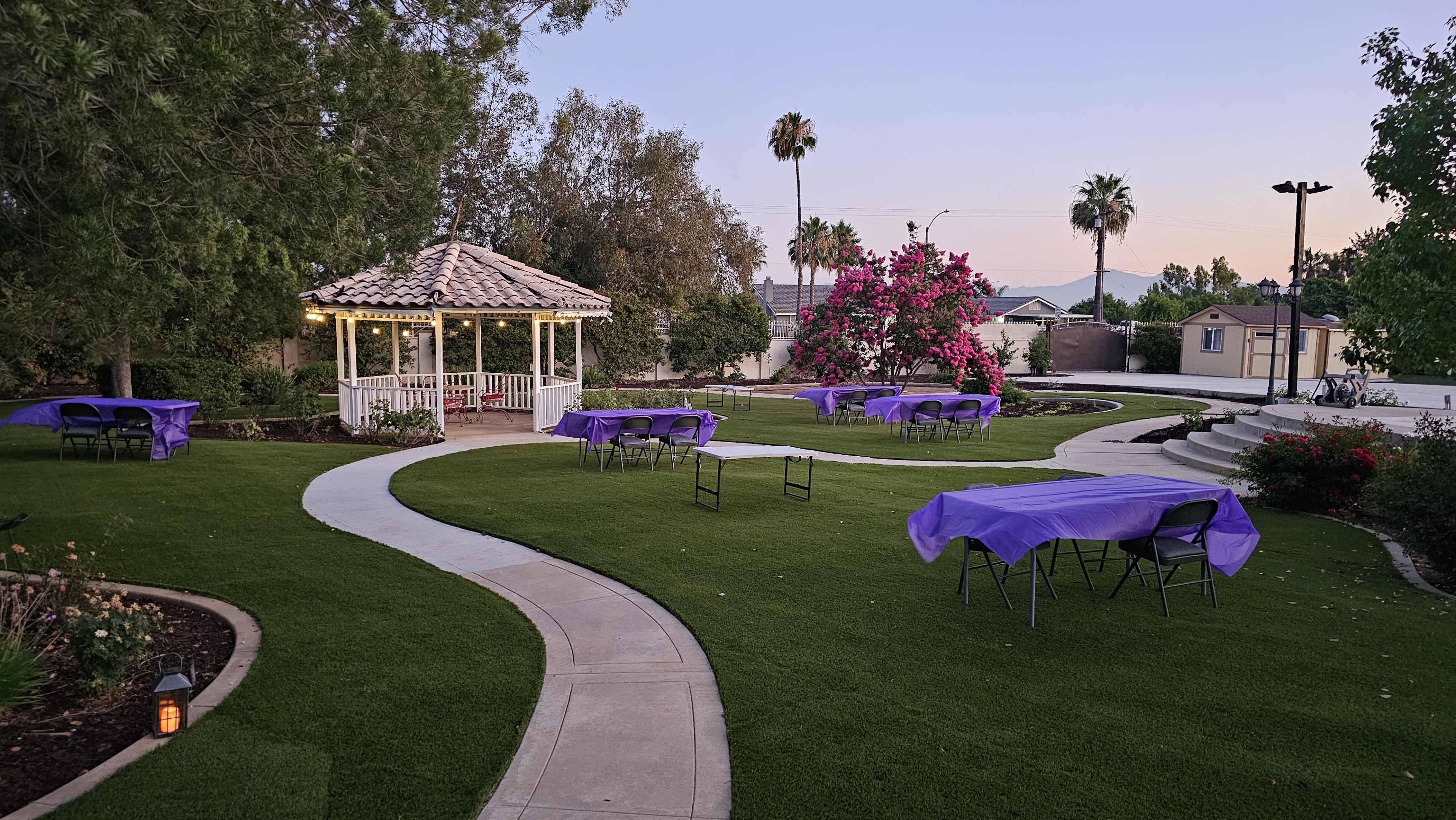 The image shows a landscaped area with a gazebo, winding pathways, and several tables covered with purple tablecloths surrounded by greenery and blooming flowers.