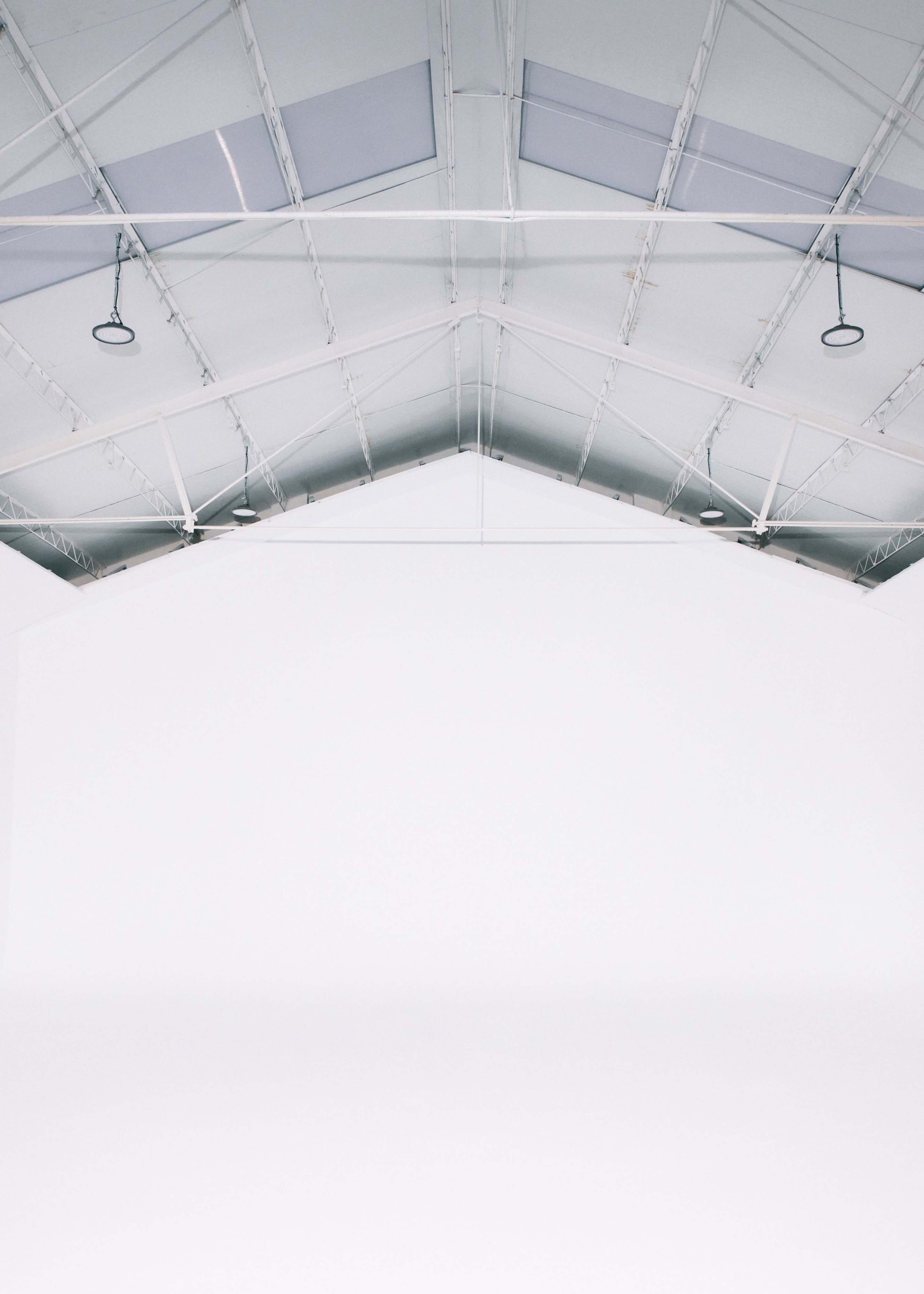 The image shows a large, empty room with a high, peaked ceiling made of metal beams and a completely white wall.