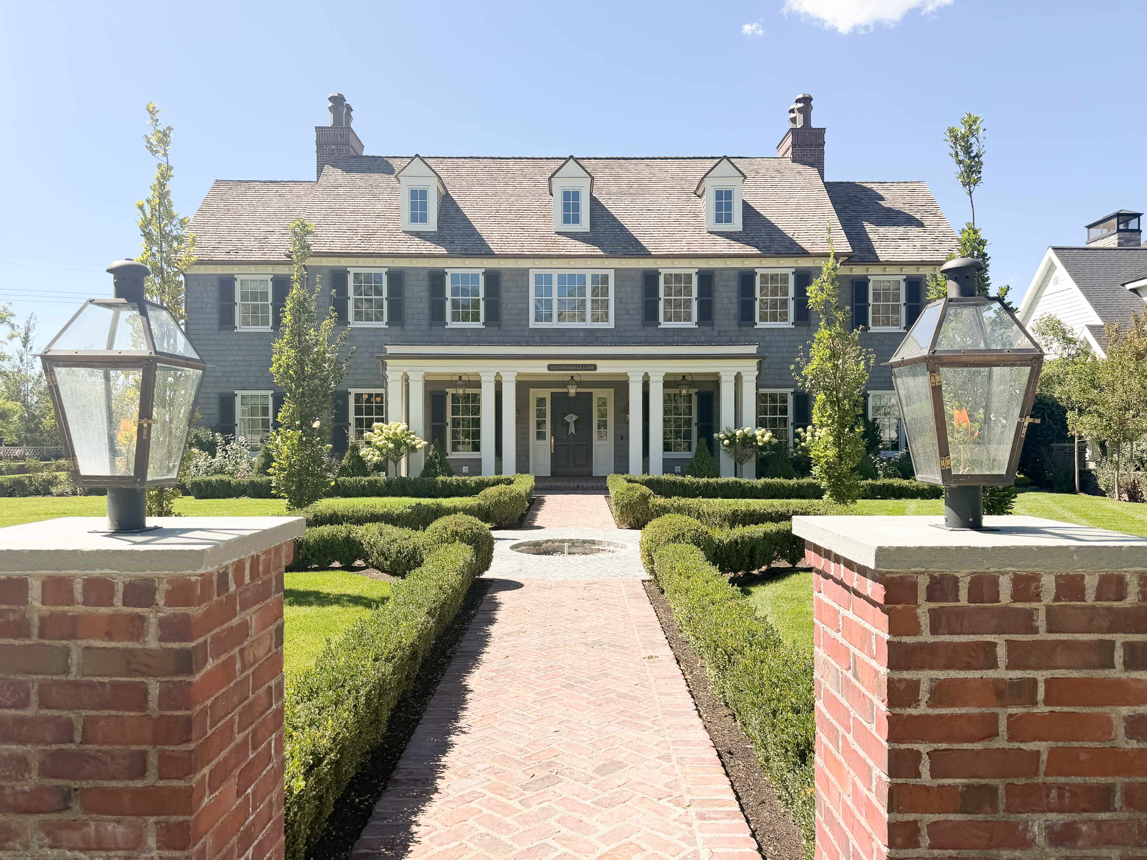 A large, two-story gray shingle house with white trim features a symmetrical facade and is surrounded by neatly manicured hedges and pathways.