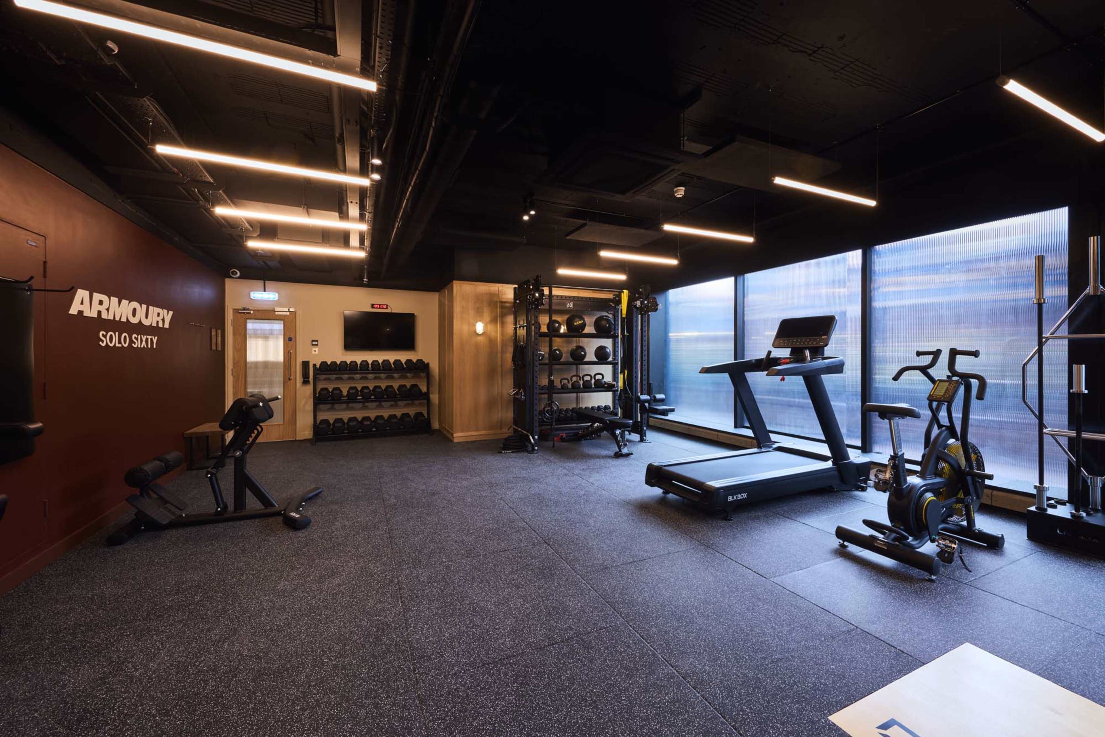 The image shows a modern gym interior featuring various exercise equipment, including a treadmill, stationary bike, weights, and a strength training station.