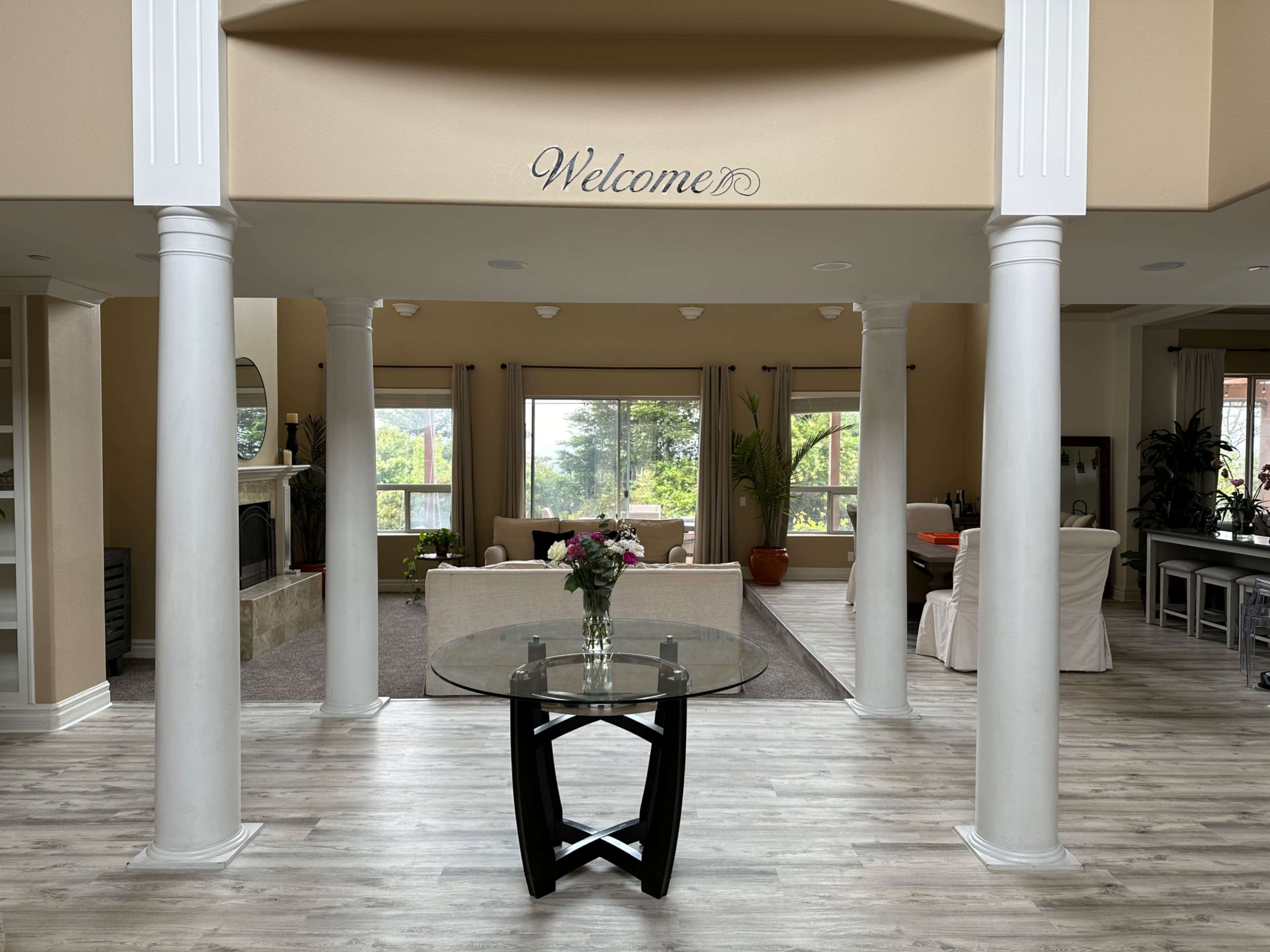 A spacious living area with decorative columns, a glass table in the foreground, and large windows framing a view of greenery outside.
