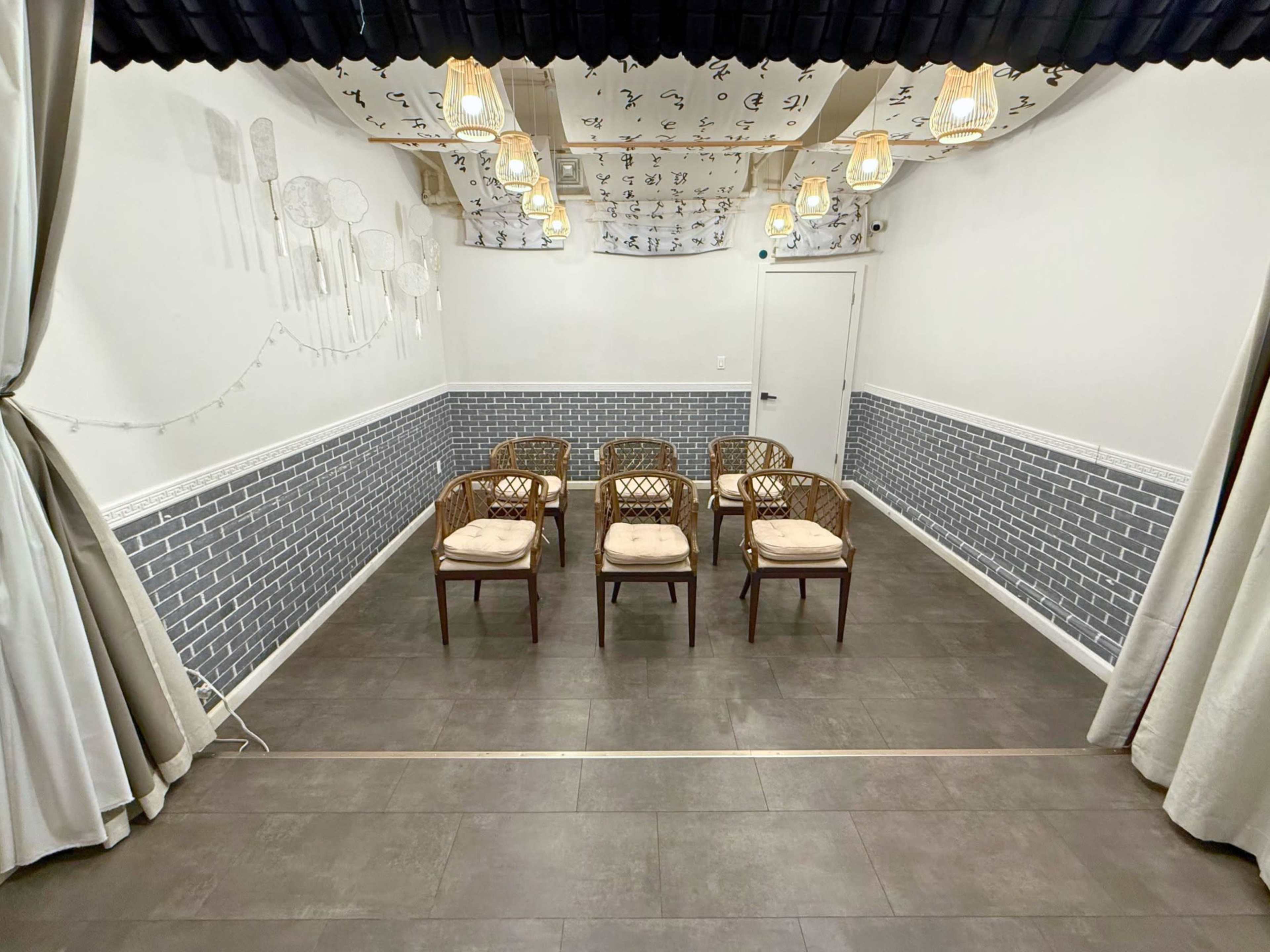The image shows a small, empty room with six wooden chairs arranged in a semicircle on a dark tile floor, and a patterned ceiling with pendant lights.