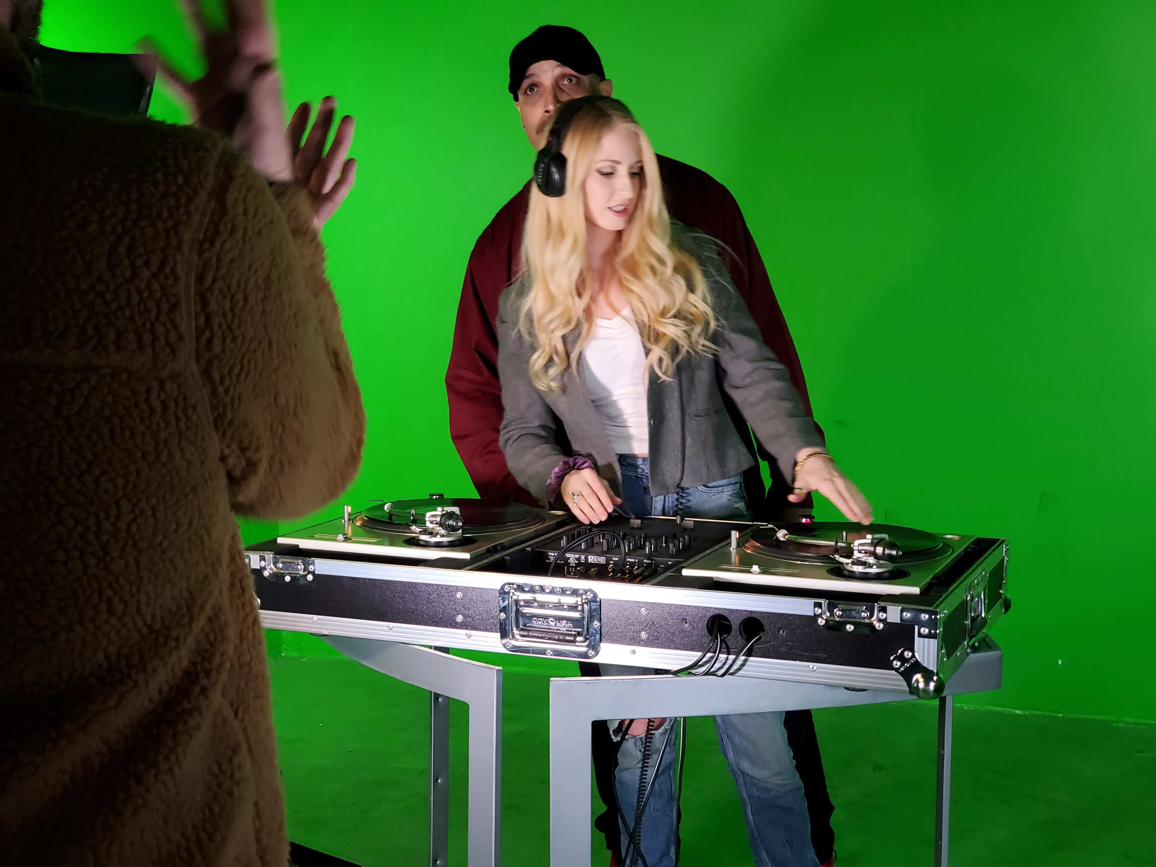 A woman stands at a DJ table with turntables, while a man behind her appears to assist or guide her, set against a green screen backdrop.