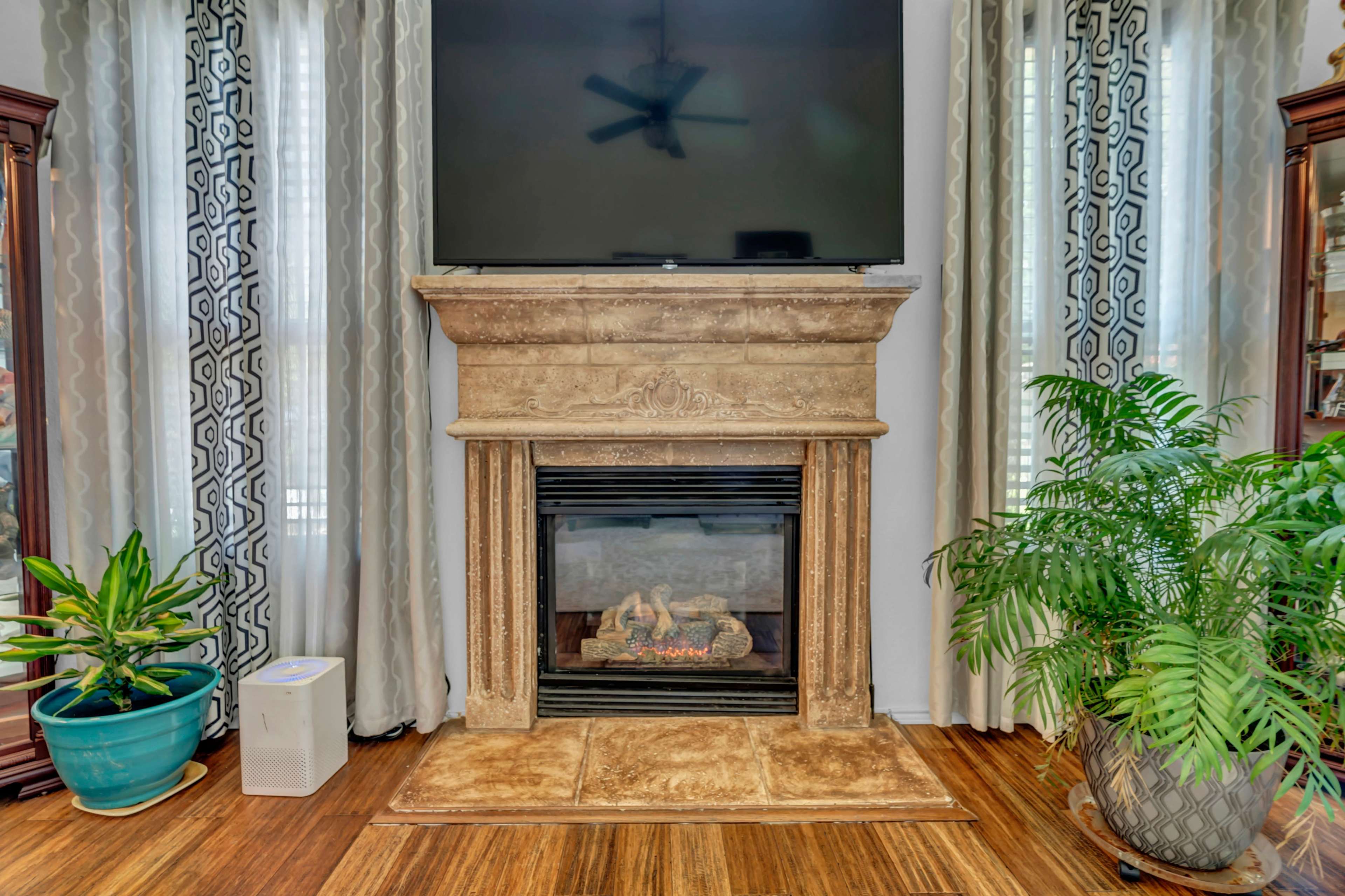A living room with a stone fireplace beneath a television, flanked by curtains, indoor plants, and wooden flooring.