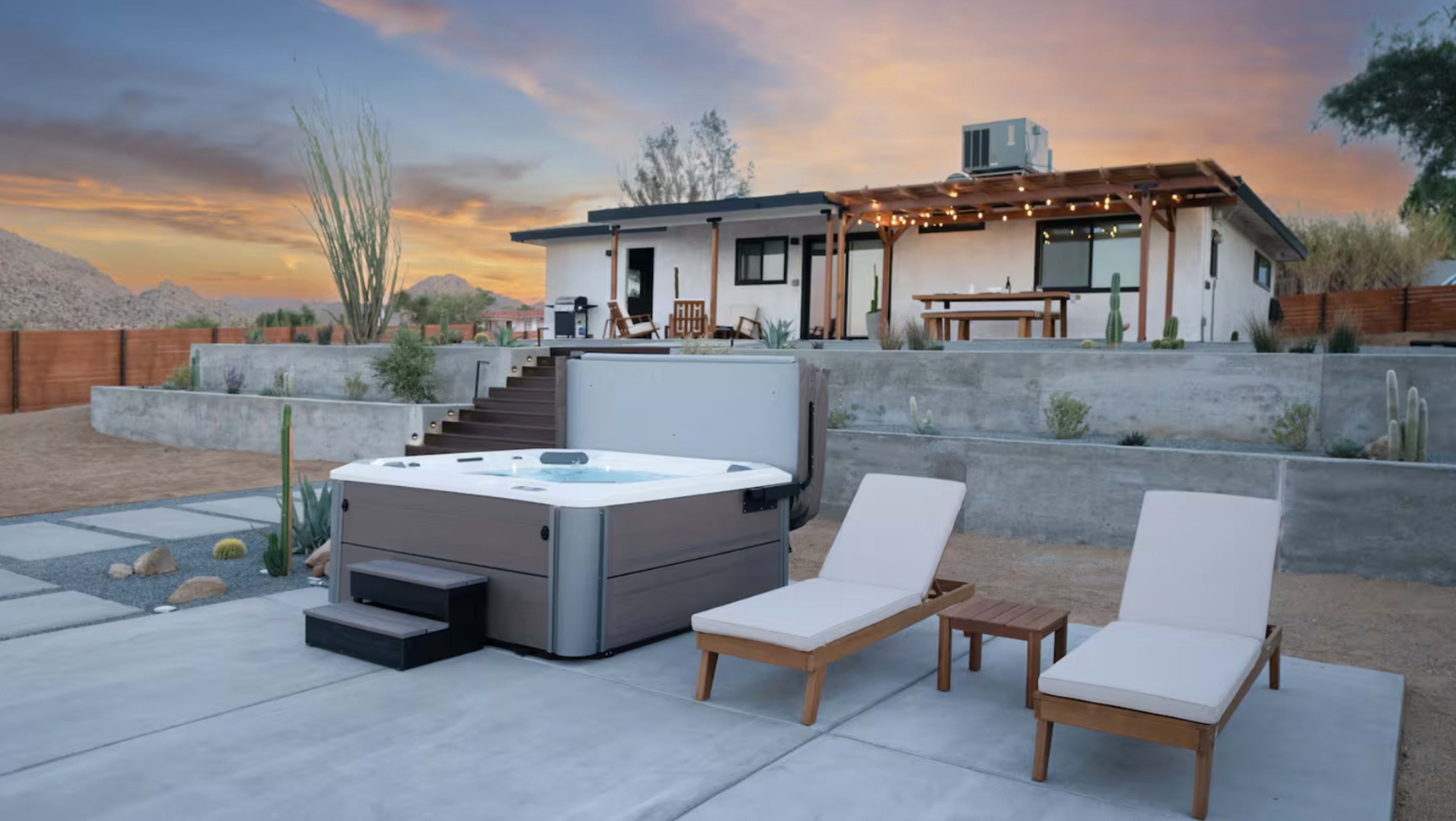 A hot tub and two lounge chairs sit on a concrete patio next to a modern house, with mountains in the background and a sunset sky.