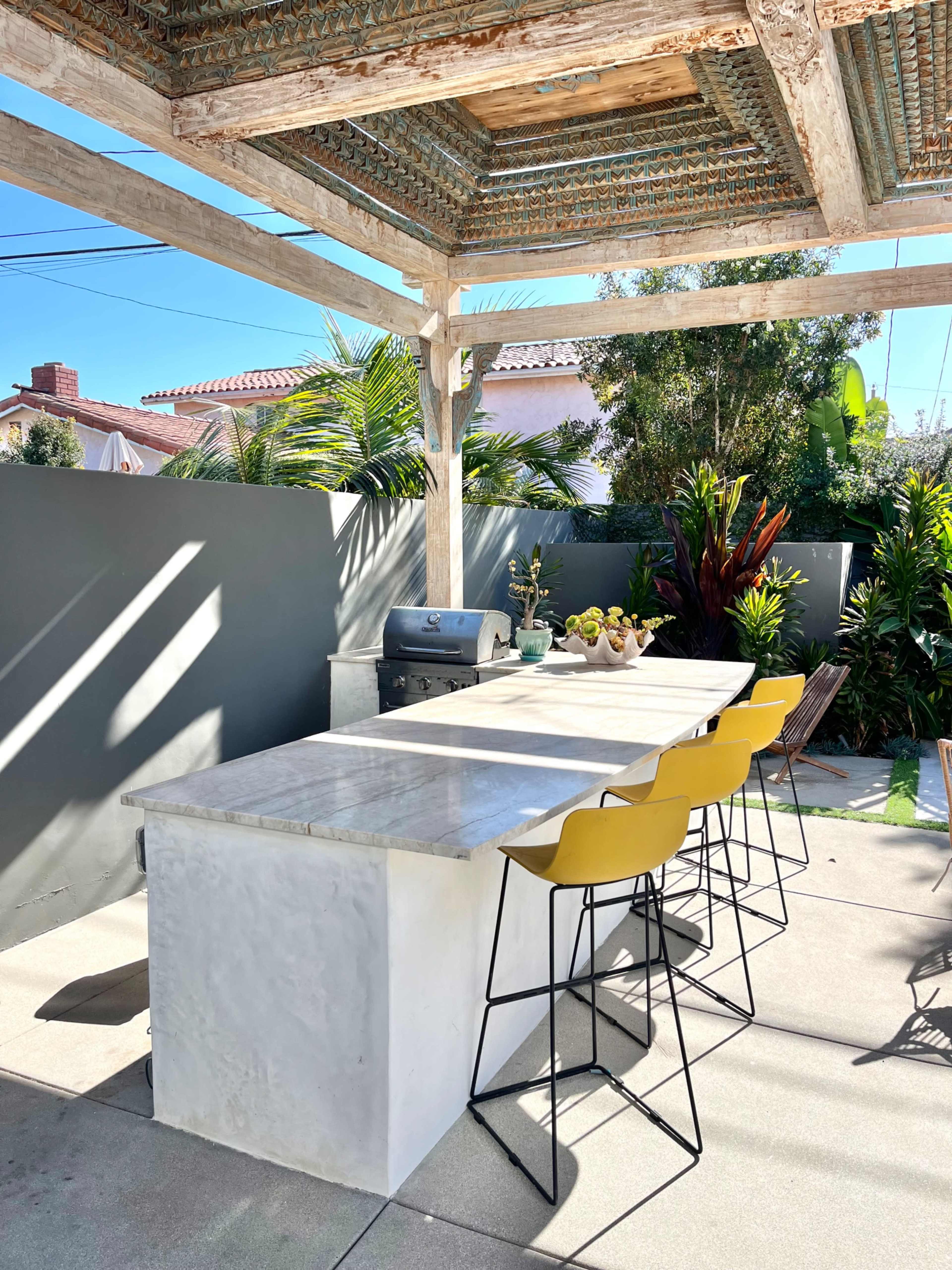 The image shows an outdoor kitchen with a marble countertop, metal bar stools, and a grill under a wooden pergola, surrounded by greenery.