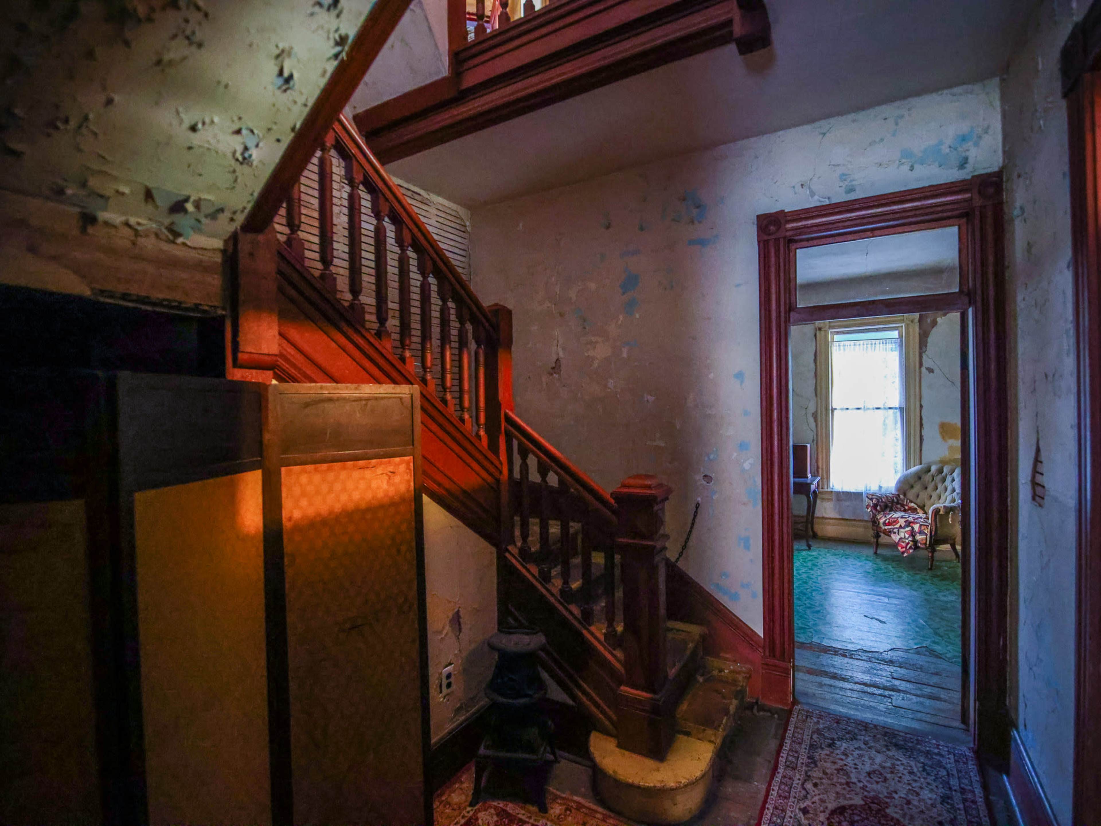 The image shows a dimly lit hallway featuring a staircase, worn walls, and a doorway leading to another room with a couch.