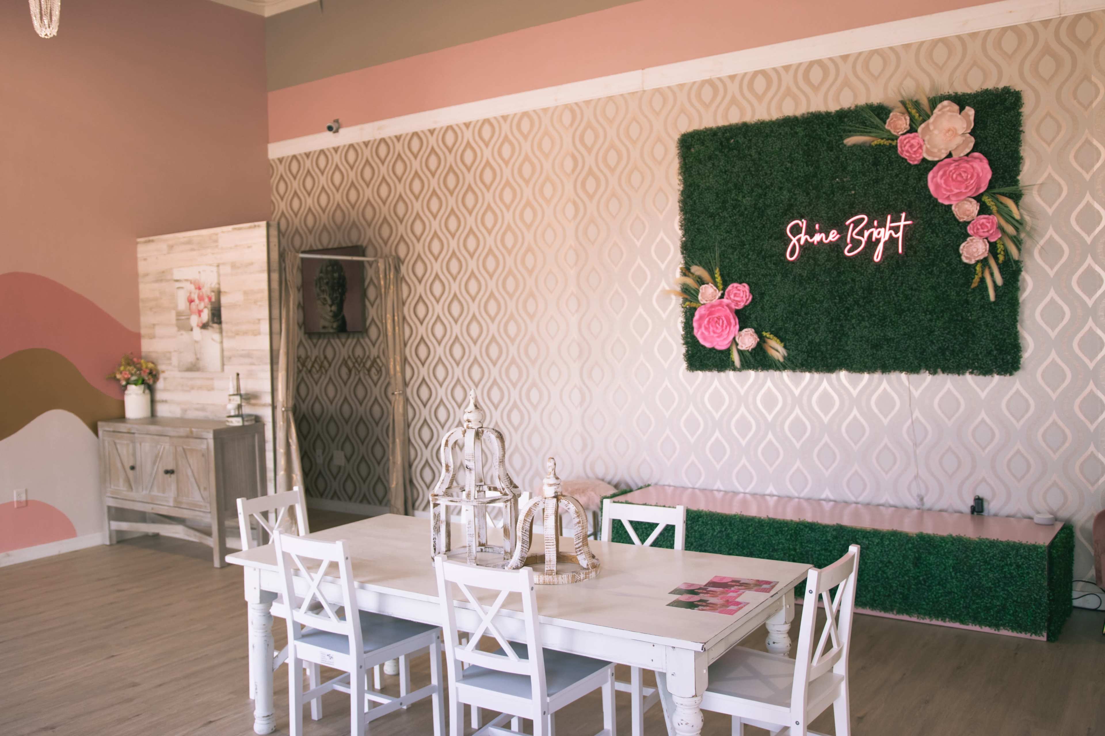 The image shows a softly decorated indoor space featuring a table with white chairs, a green wall with pink flowers, and a neon sign that reads "Shine Bright."