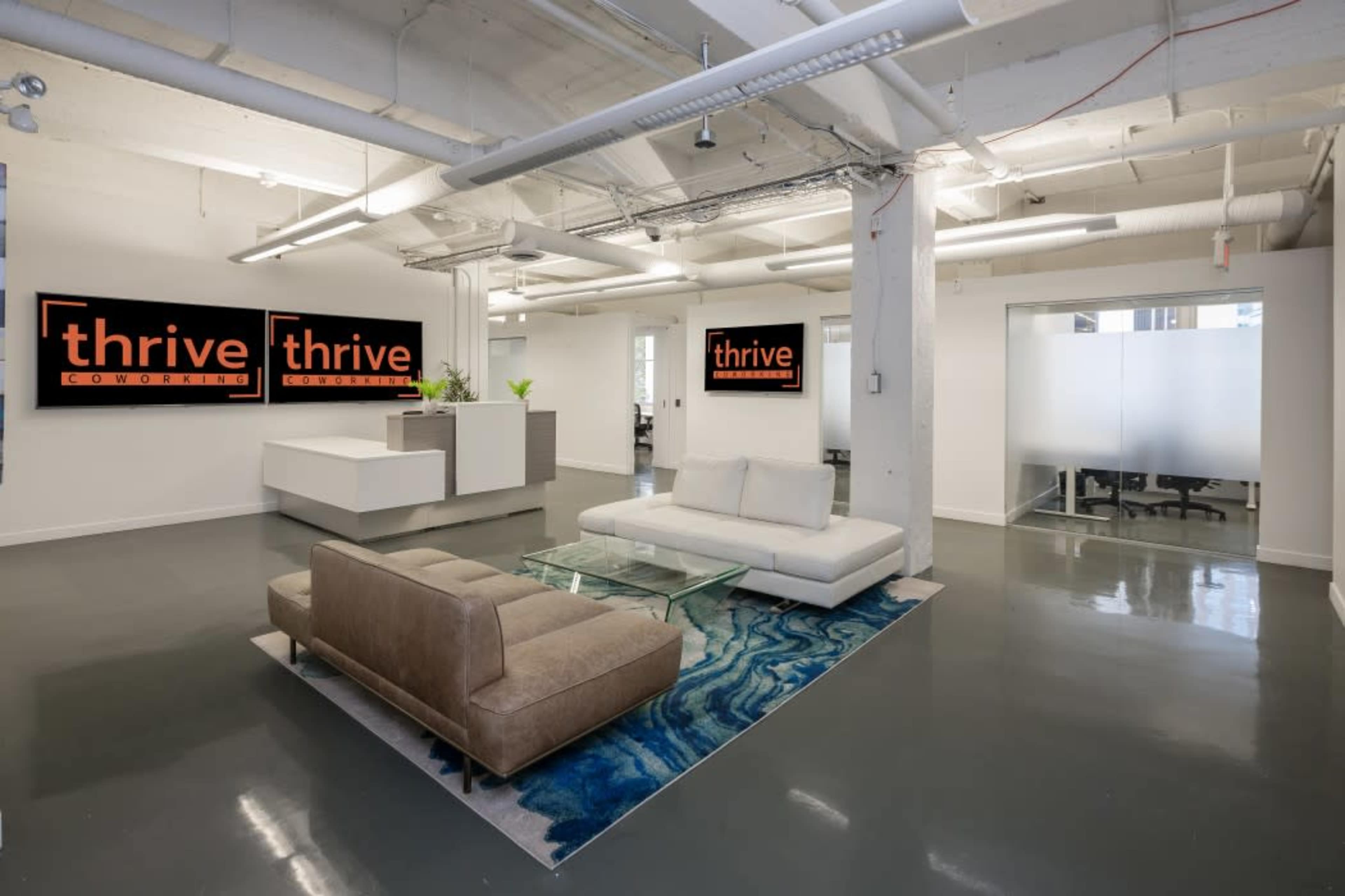 A modern coworking space with a seating area, featuring a white sofa and a brown couch on a patterned rug, alongside bold "thrive" signage on the walls.