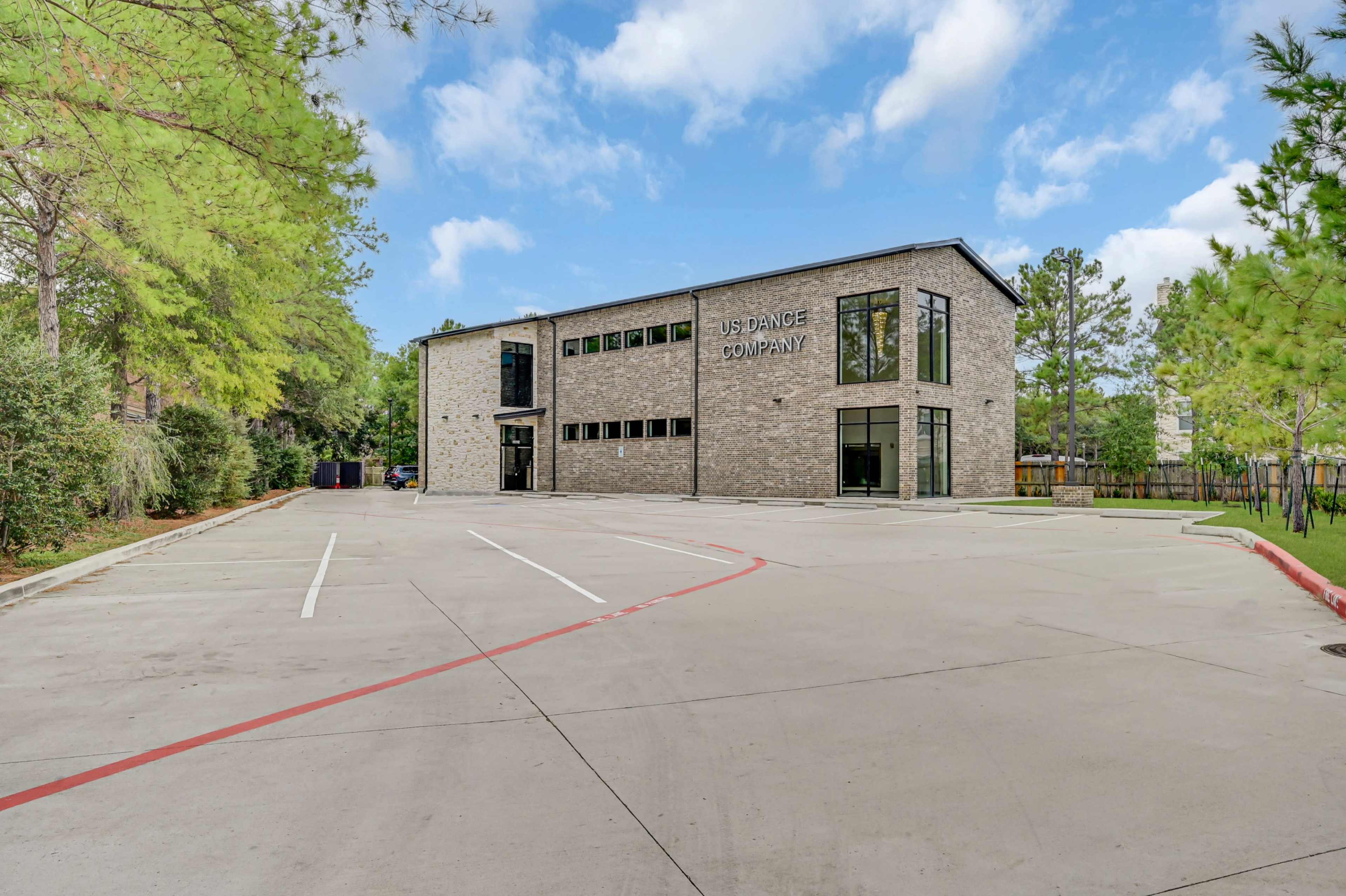 The image shows a modern stone building with large windows, labeled "US Dance Company," set against a backdrop of green trees and a clear sky, with a mostly empty parking lot in front.