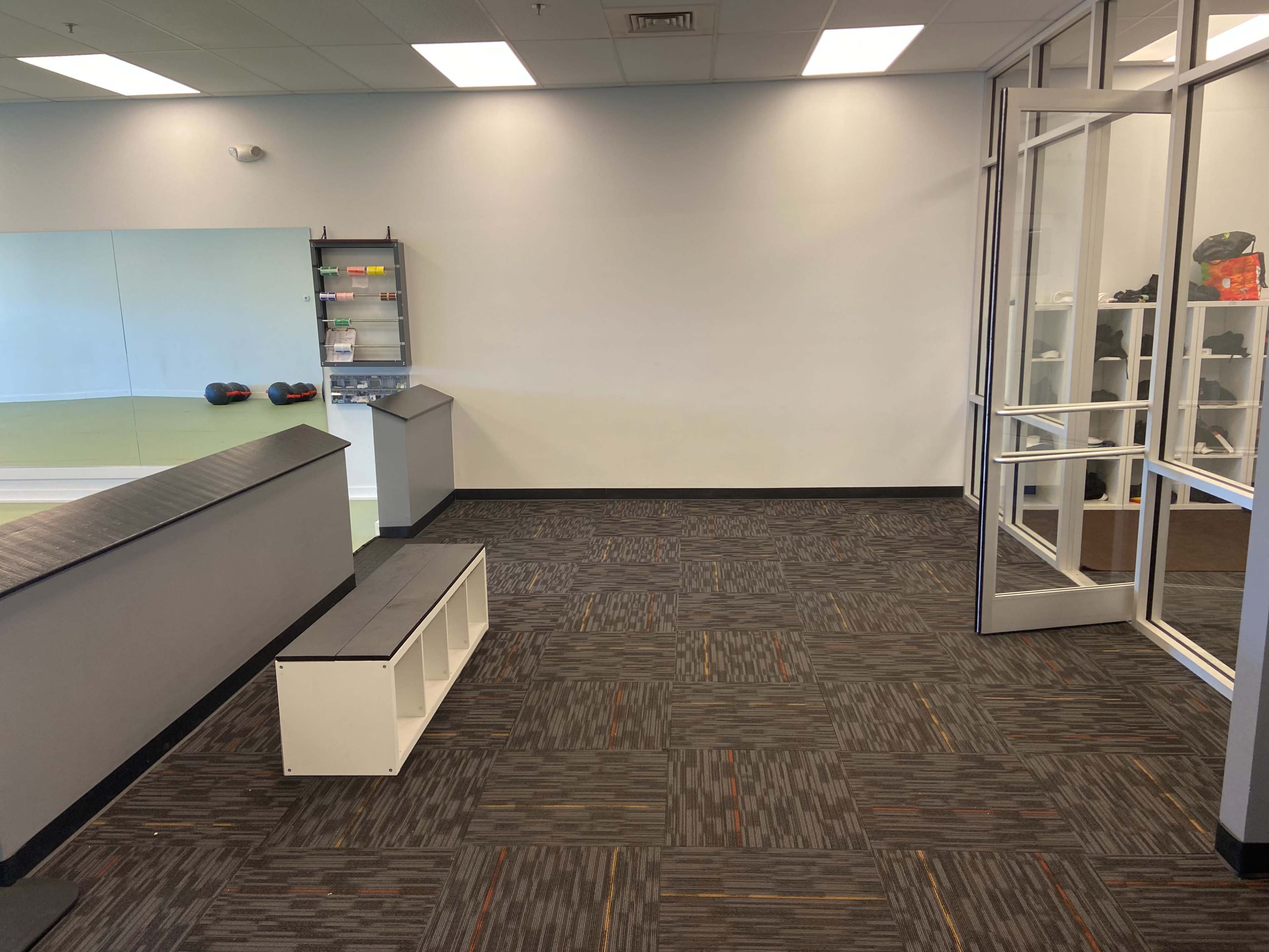 A spacious area with a gray and black carpet, a low white bench, and large windows leading to an outside entrance, alongside a wall with fitness equipment on display.
