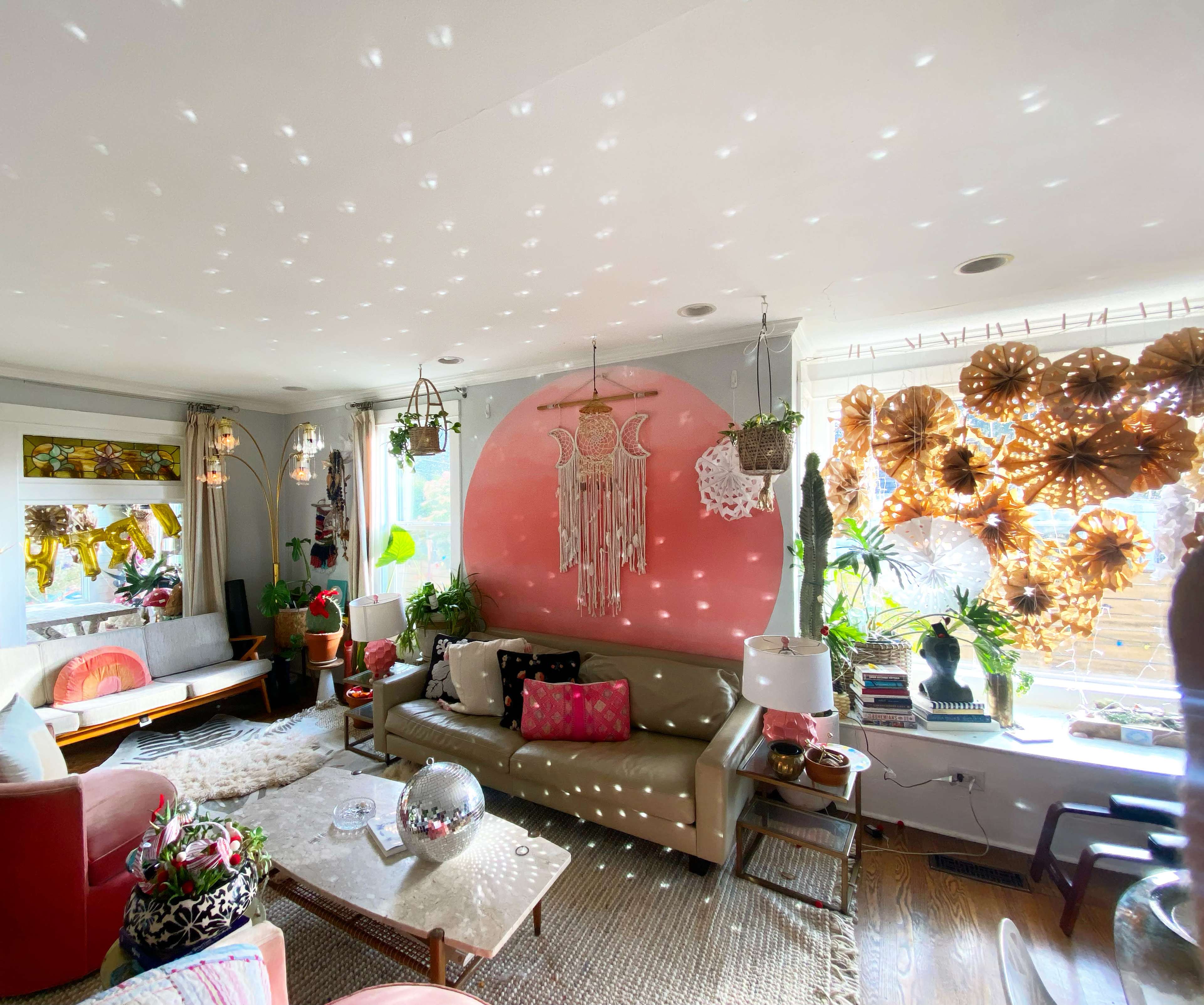The living room features a mix of modern furniture and decorative plants, with a large pink circular wall art piece and colorful light reflections on the ceiling.