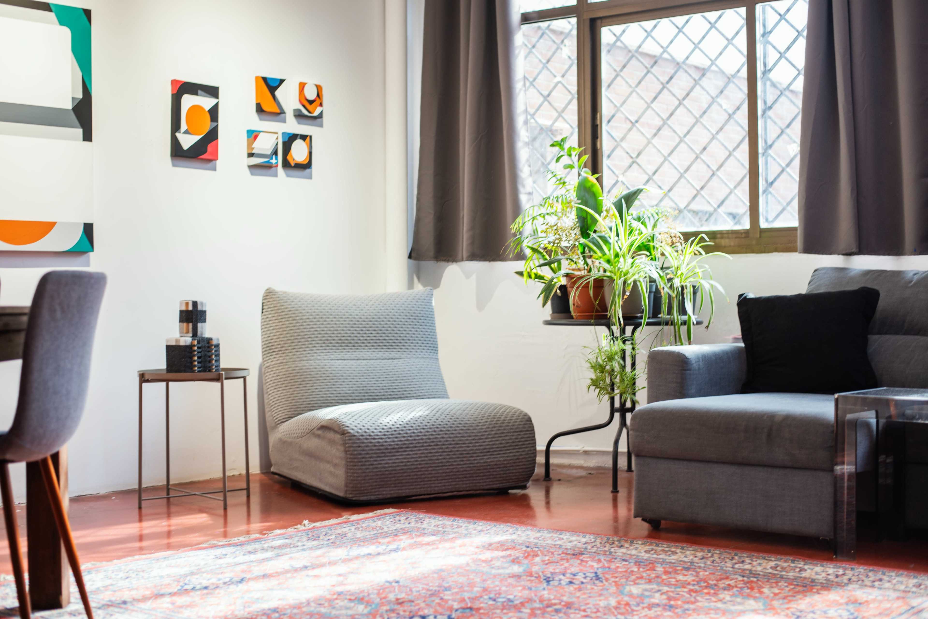 The image shows a modern living room with a plush gray chair, a sectional sofa, a potted plant near a window, and abstract wall art.