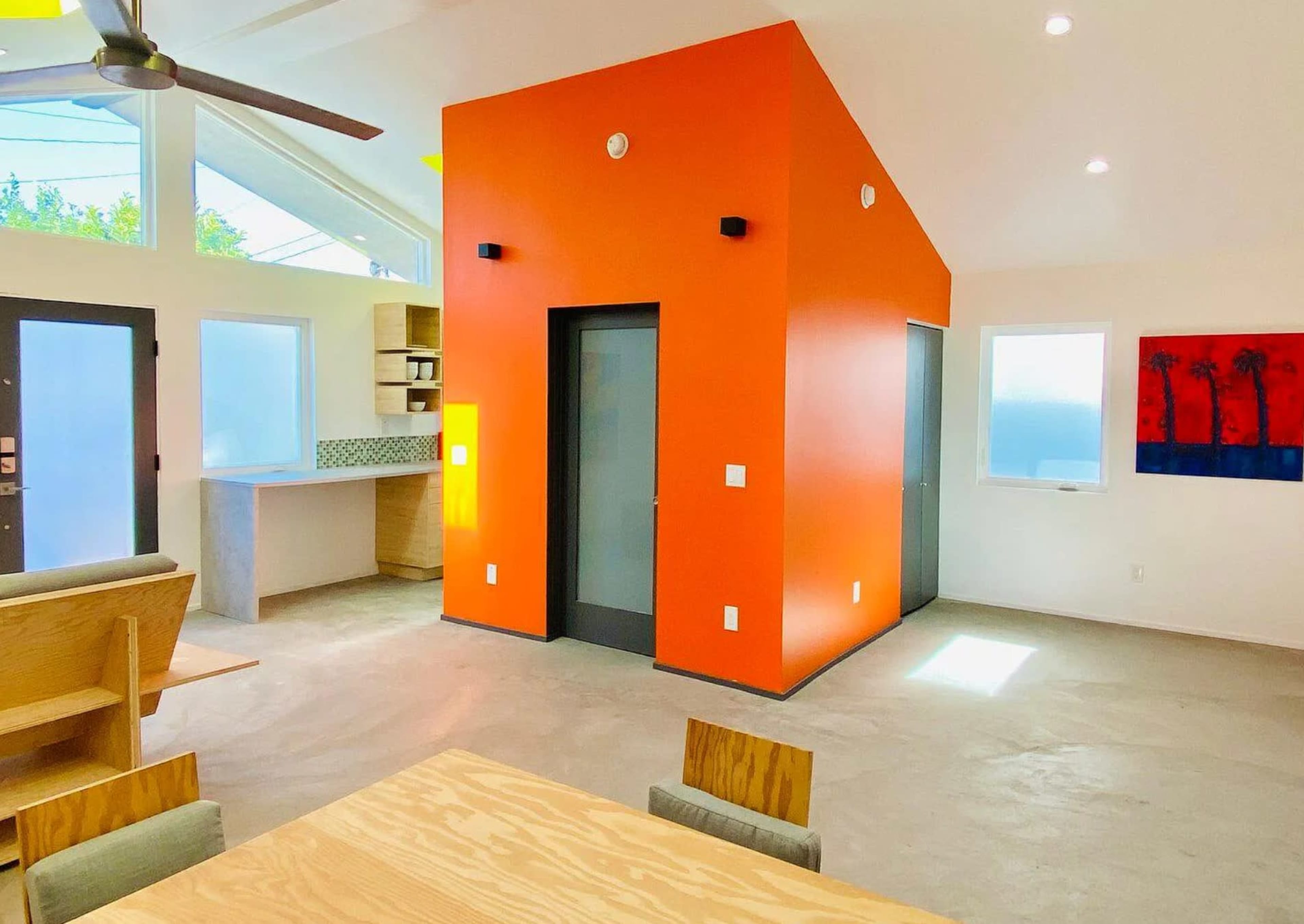 A modern, open-concept living space featuring a striking orange wall, concrete flooring, and large windows with a view.