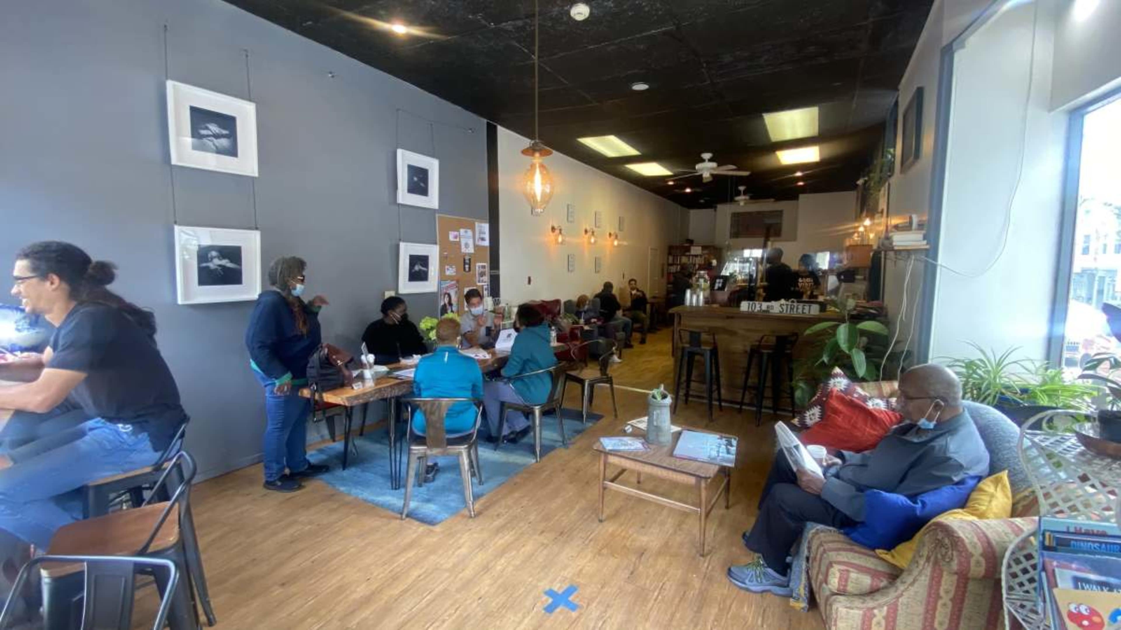 A busy coffee shop features a mix of patrons seated at tables and couches, with artwork displayed on the walls and natural light streaming through large windows.