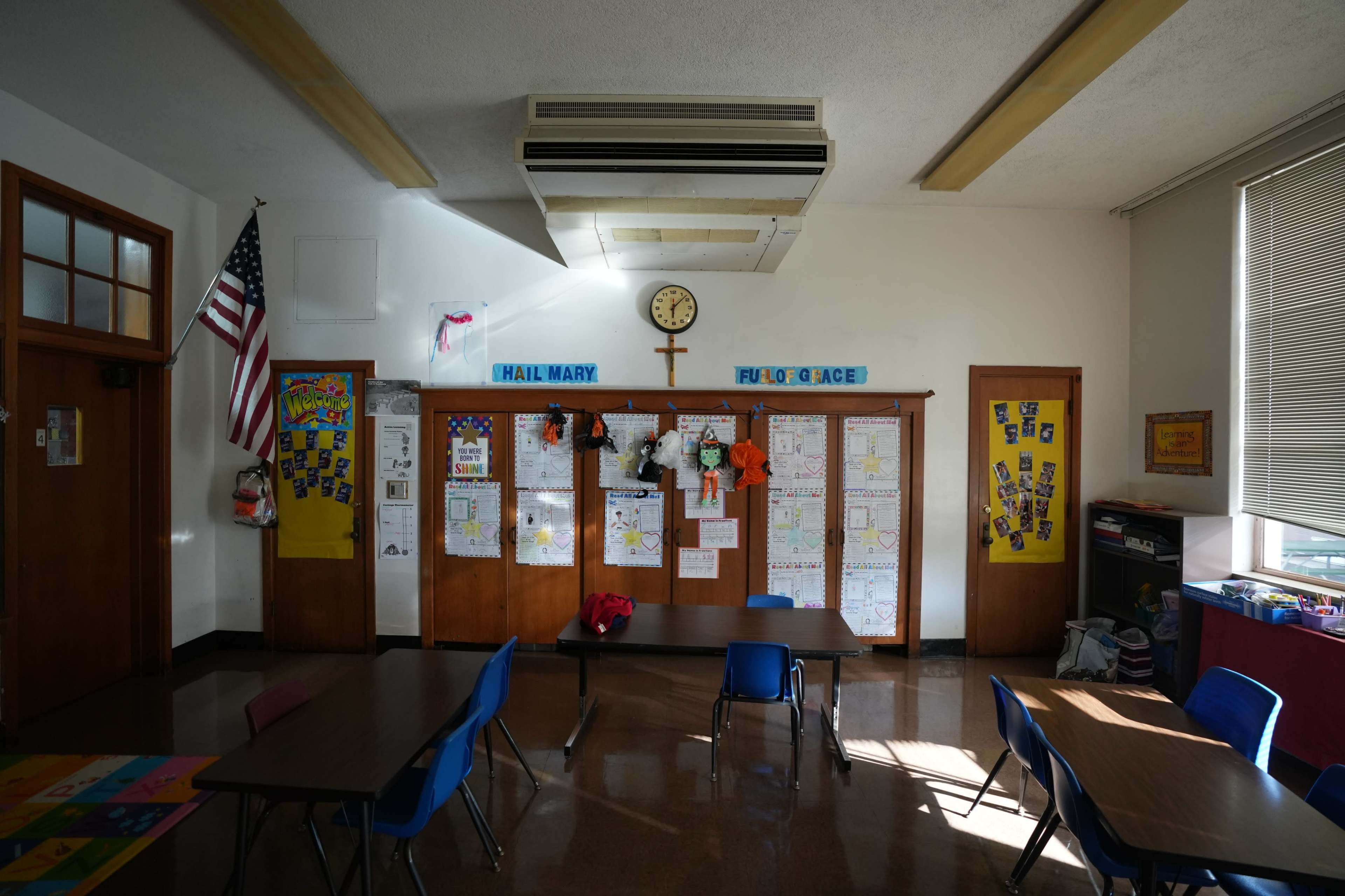 A classroom features blue chairs and wooden bulletin boards displaying various papers and artwork, along with a clock and an American flag on the wall.