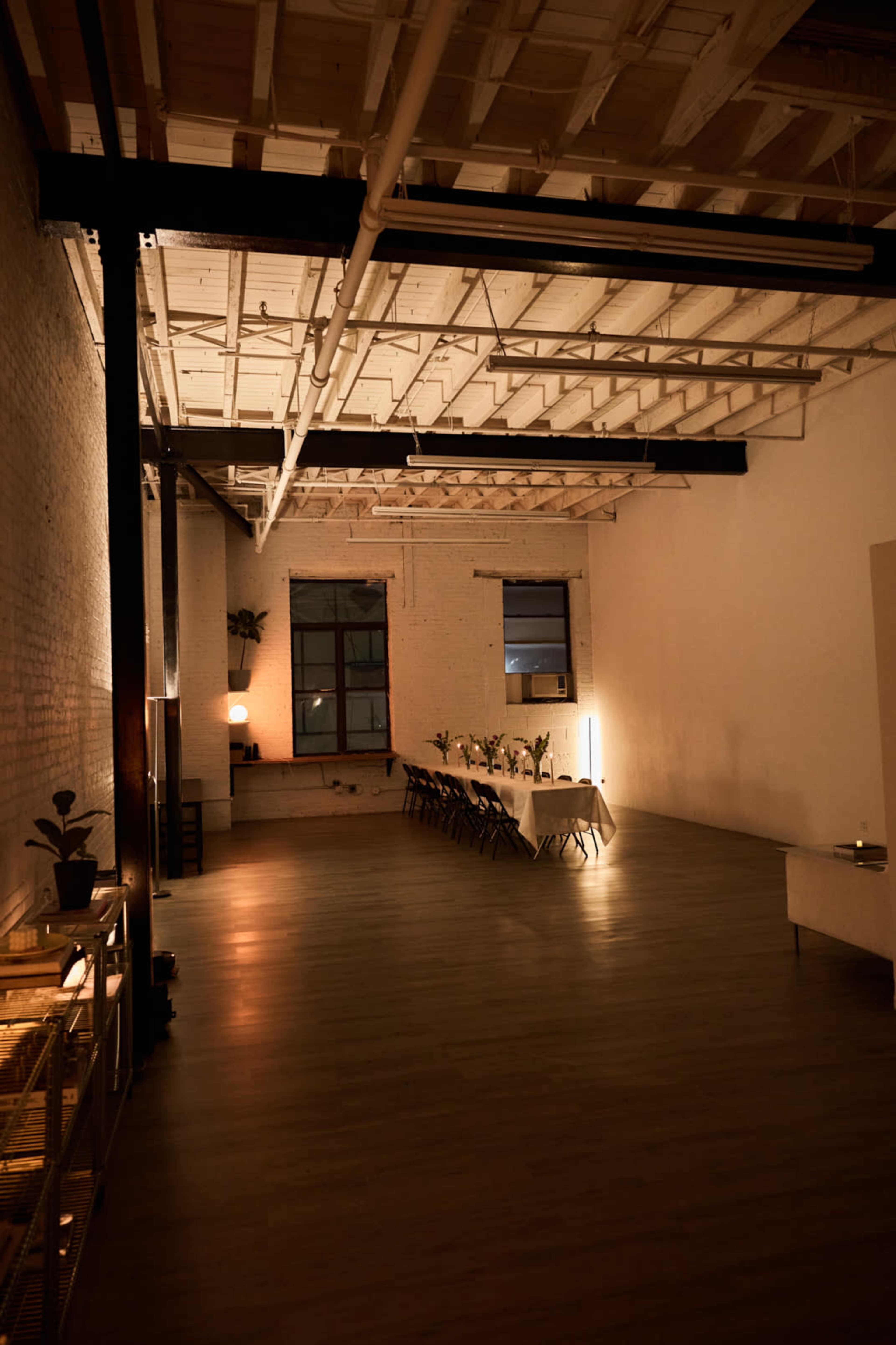 A spacious, well-lit room with wooden flooring features a long table set for a gathering, and plants are arranged along the walls.
