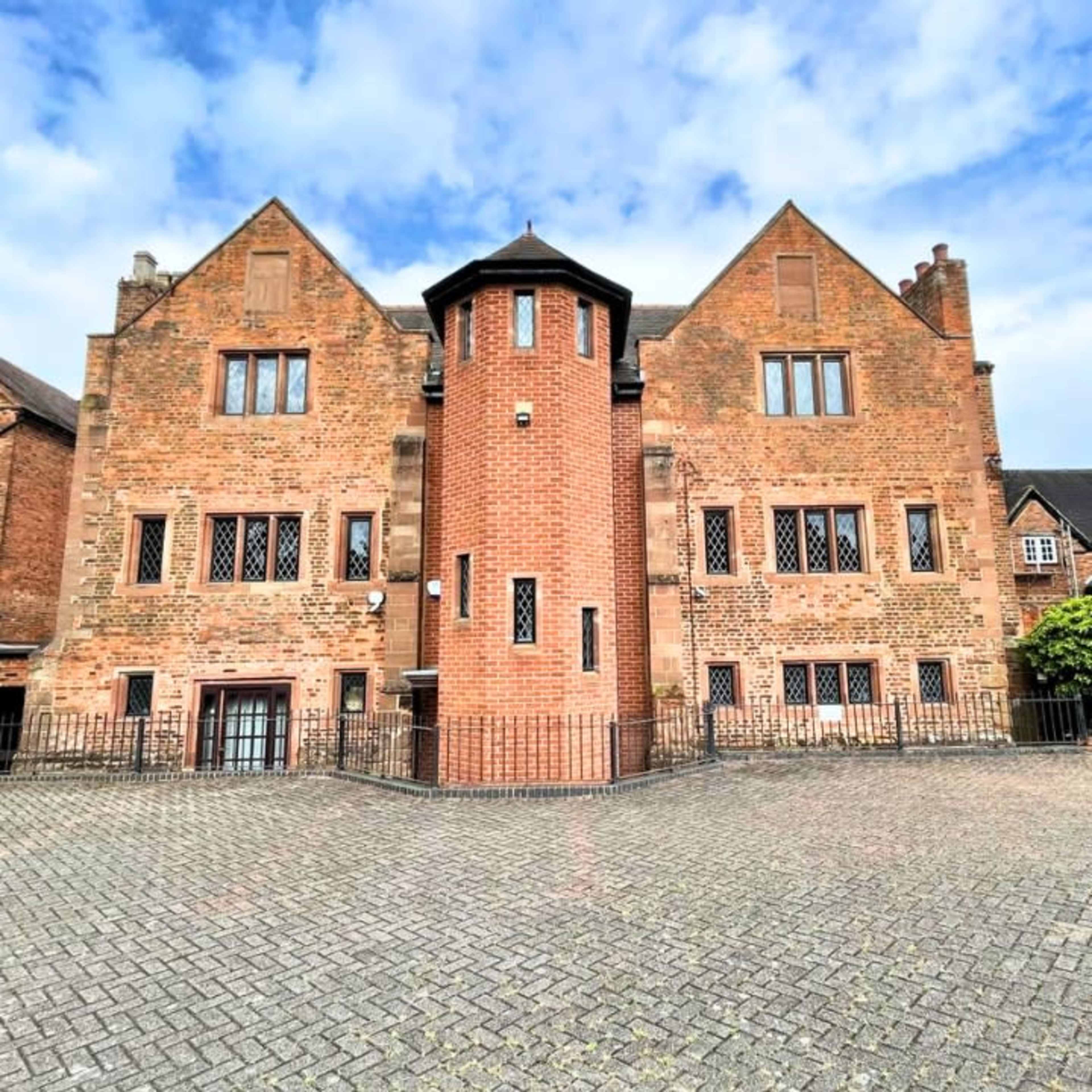 The image shows the rear facade of a brick building featuring a circular tower and multiple windows in a landscaped outdoor area.