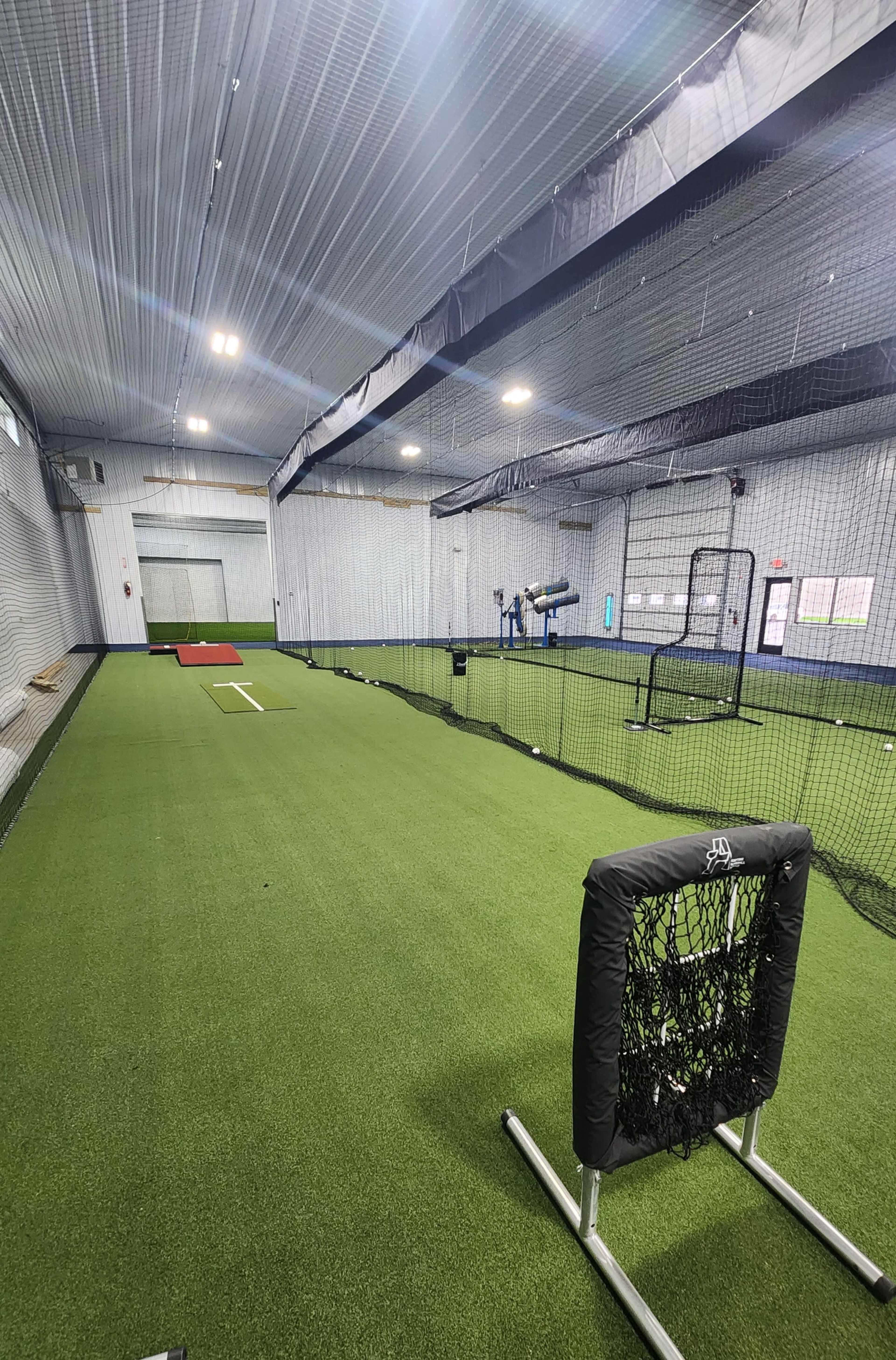 The image shows an indoor baseball training facility with artificial turf, batting cages, and equipment arranged along the space.