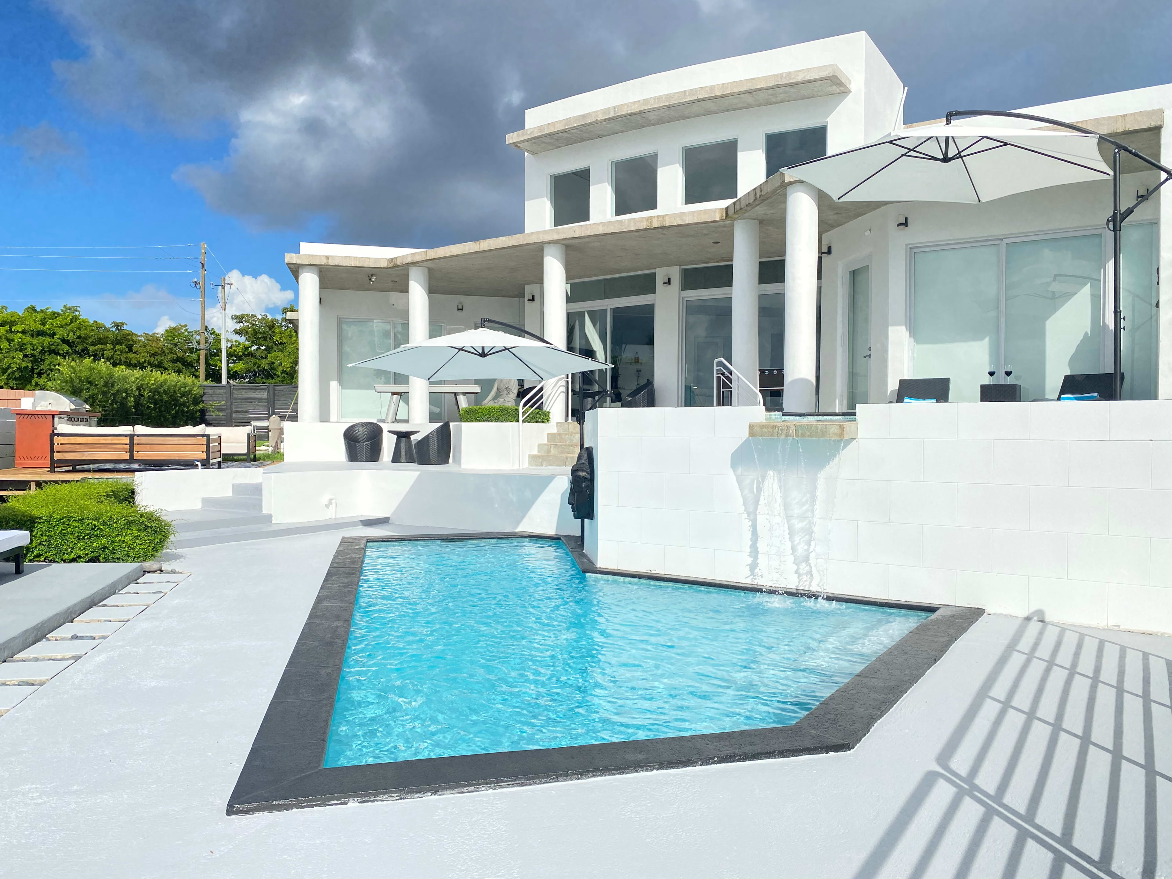 A modern house features a swimming pool with a waterfall, surrounded by outdoor seating and large umbrellas.