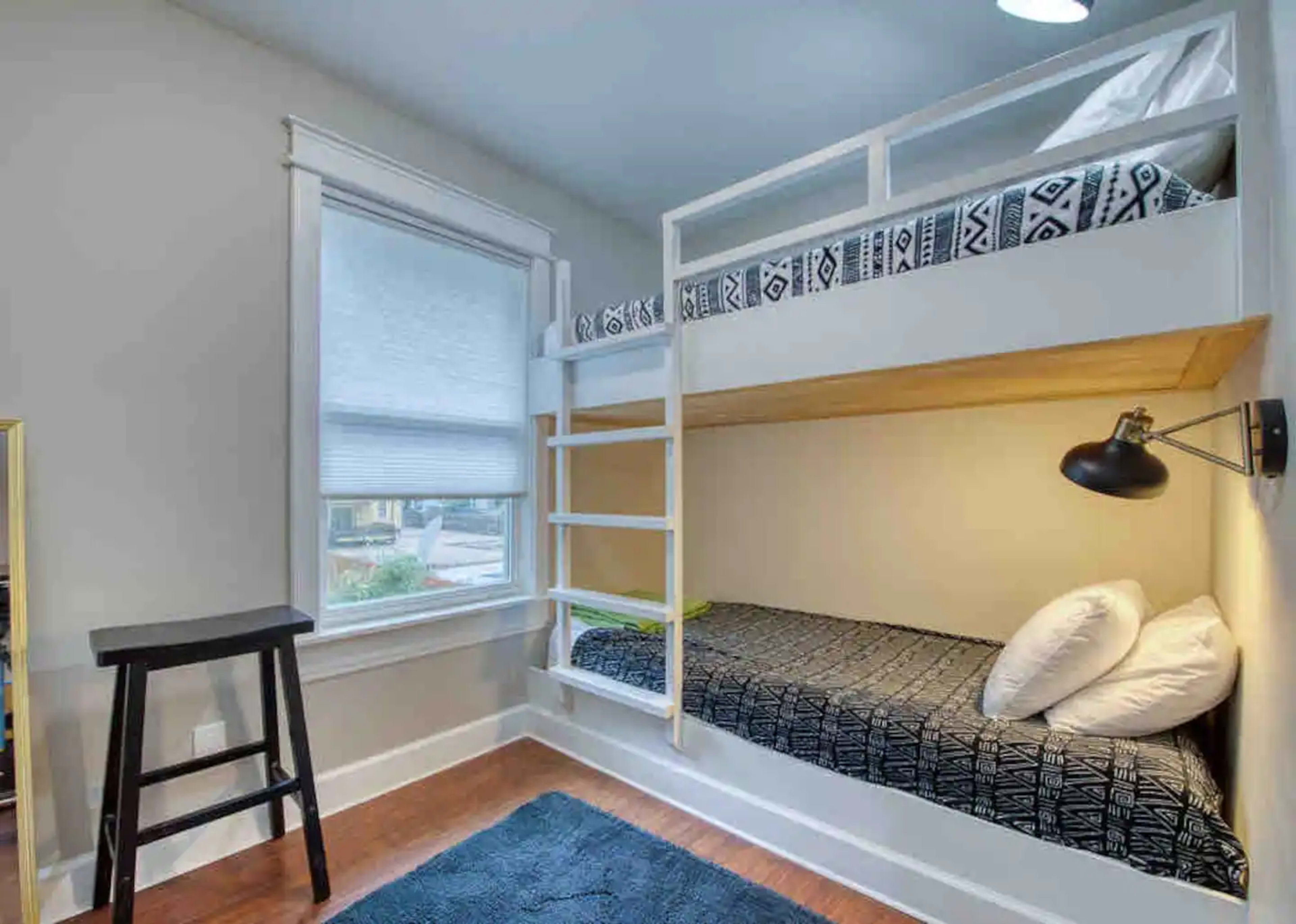 A small bedroom featuring a bunk bed with a top and bottom sleeping area, a ladder, a reading lamp, and a stool beside it.