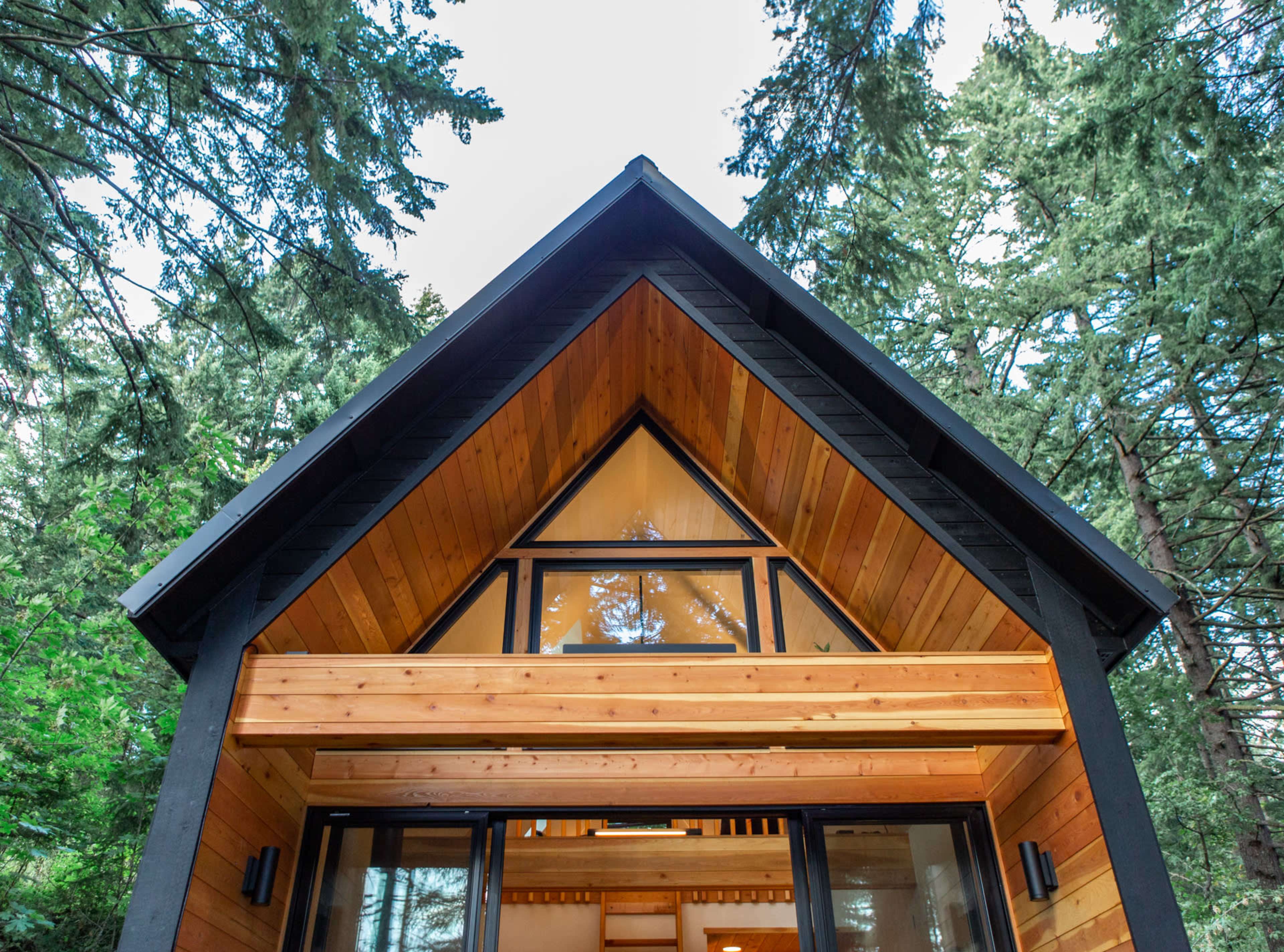 The exterior view of a wooden cabin with a triangular roof, framed by tall trees.
