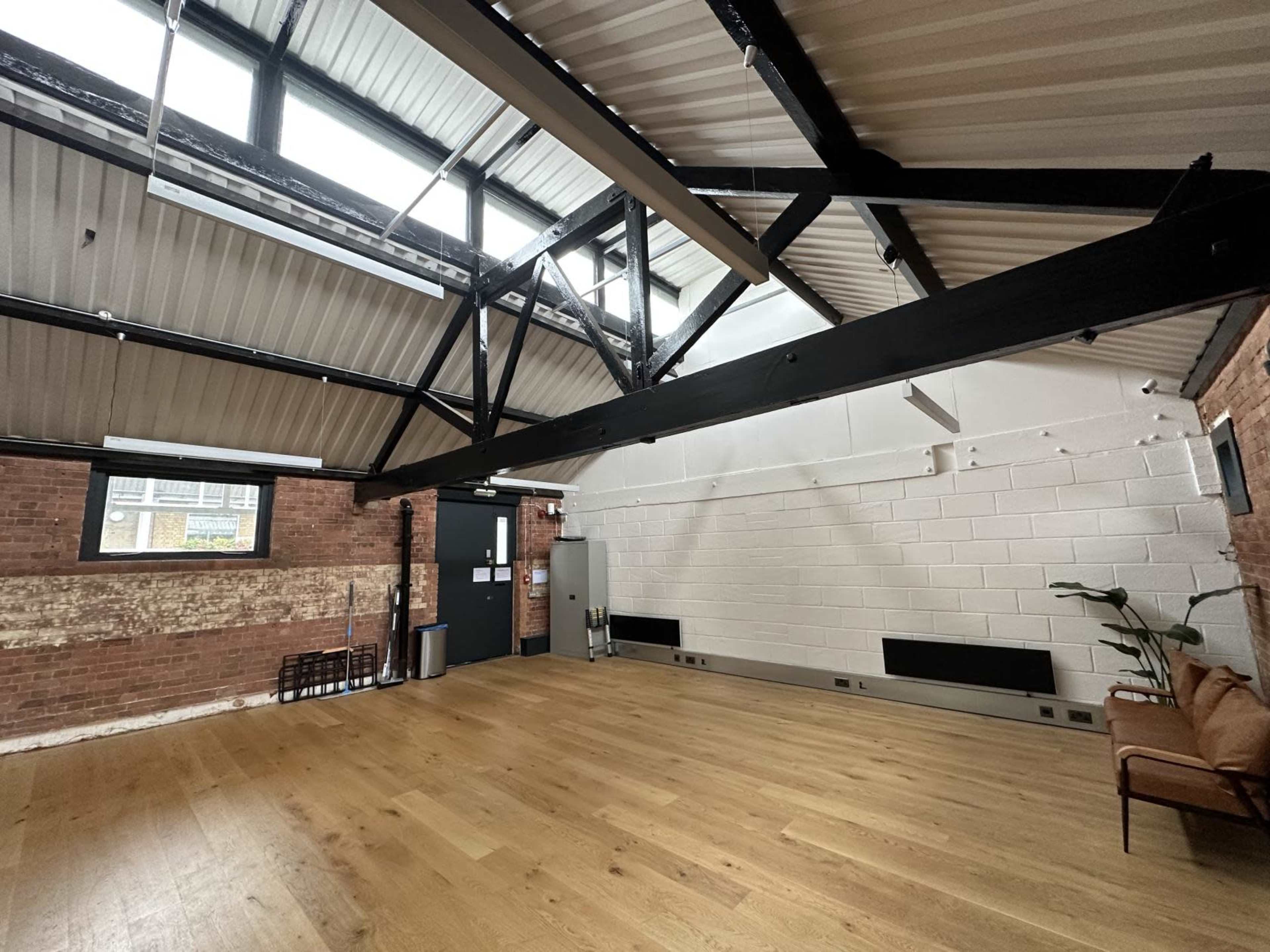 The image shows a spacious, industrial-style interior with exposed brick walls, large wooden floors, and a high ceiling featuring metal beams.